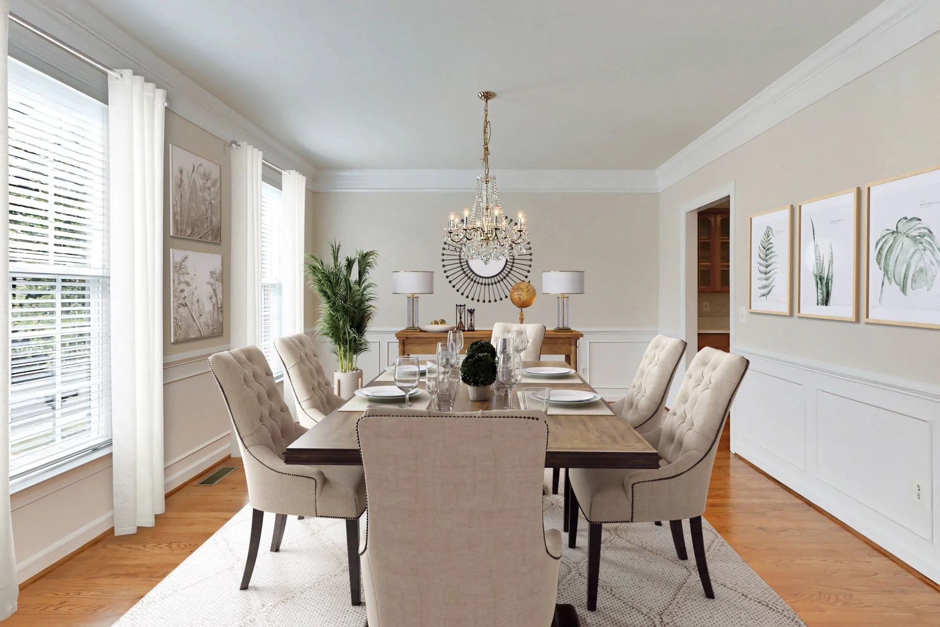 Dining room with a wooden table, eight beige upholstered chairs, a chandelier, a sideboard with lamps and decorative objects, framed botanical prints, and large windows with white curtains.