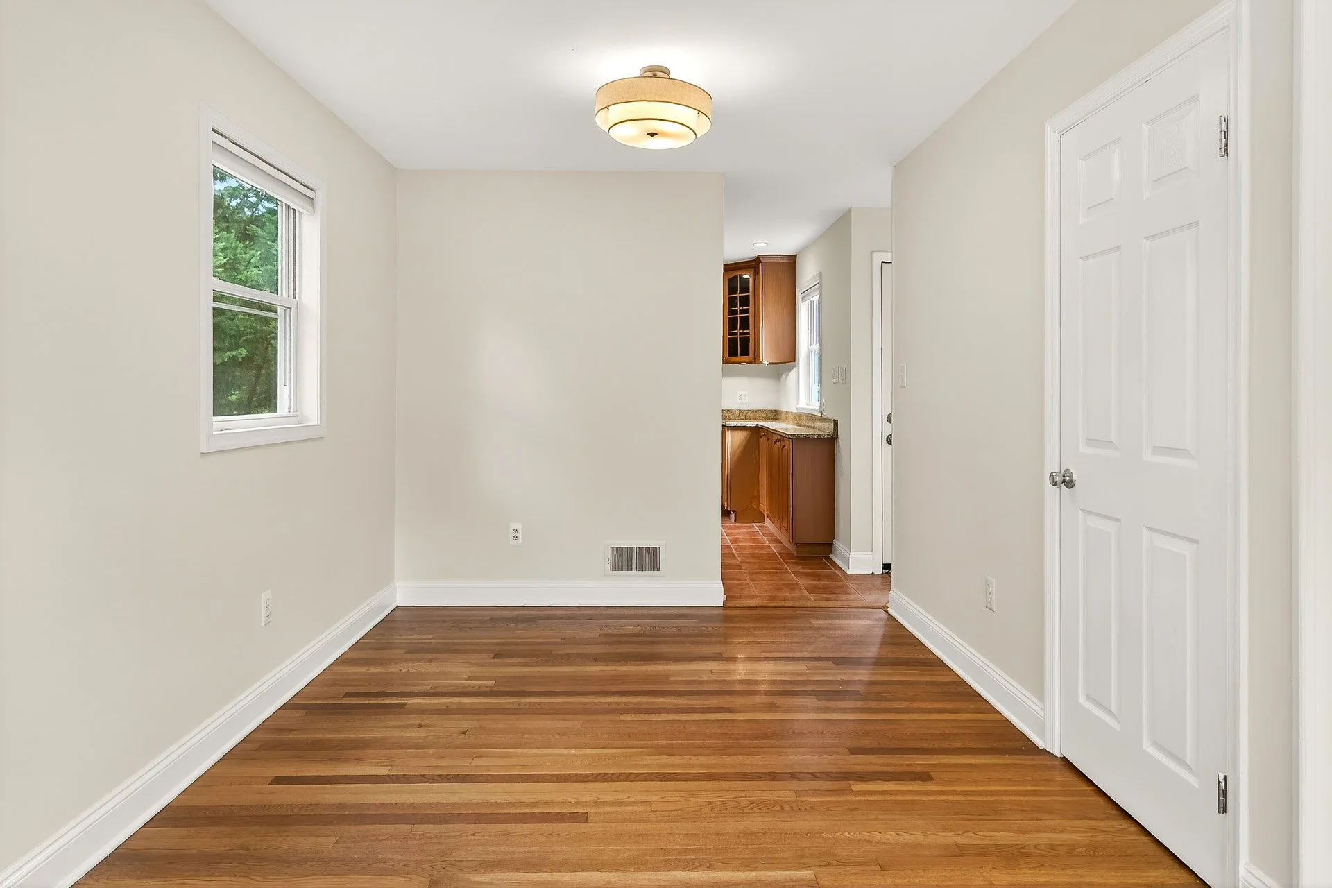 Empty living space with hardwood floors, white walls, windows, and a view into a kitchen with wooden cabinets and tiled floor.