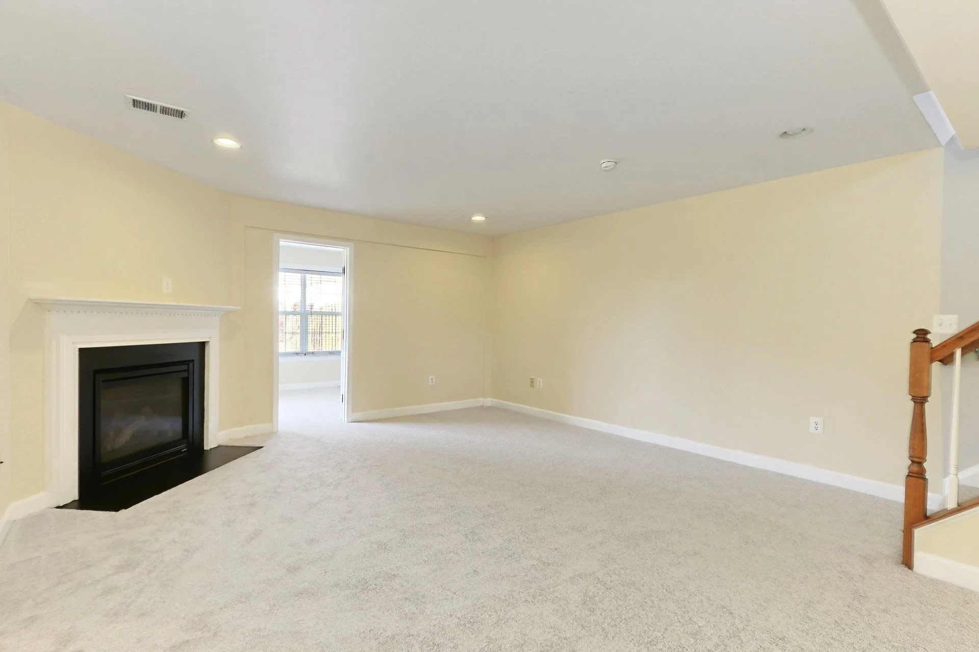 Empty living room with a fireplace, beige walls, carpeted floor, and a doorway leading to a room with a large window.