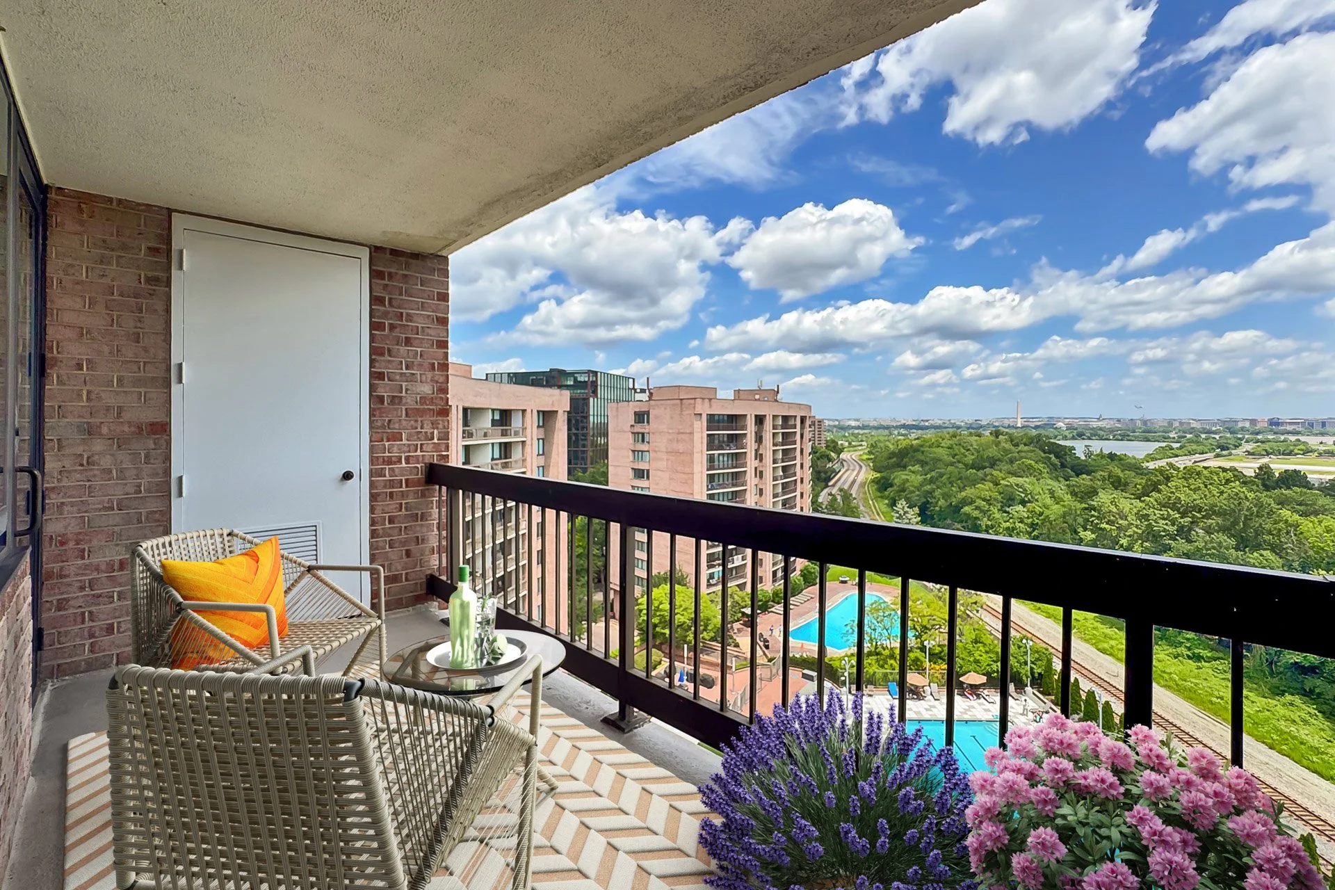 Balcony with outdoor furniture, potted flowers, and a scenic view of a cityscape, green trees, swimming pools, and a river under a partly cloudy sky.