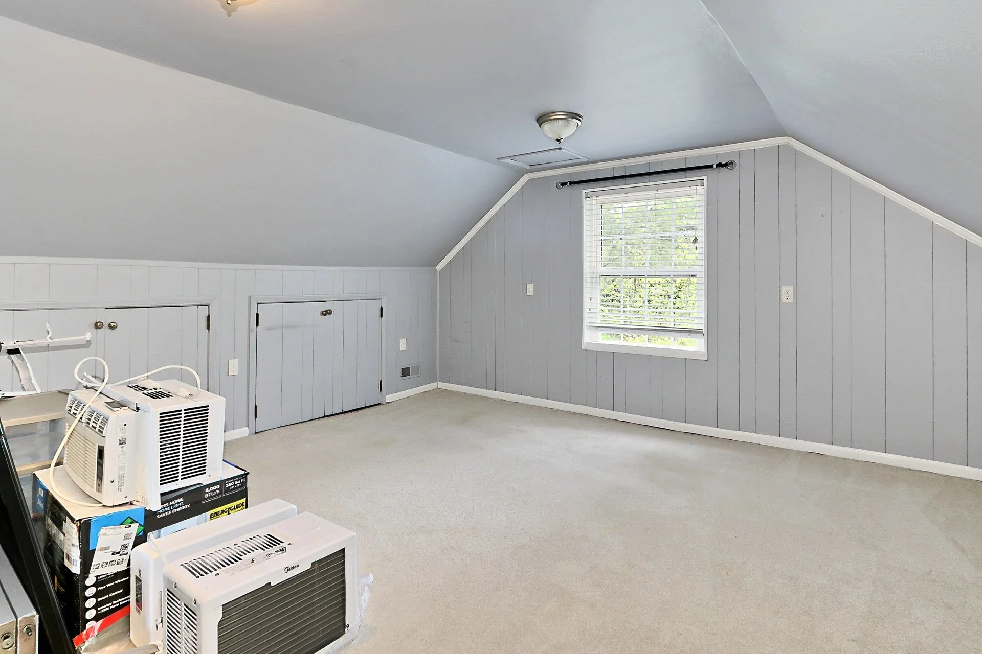 Empty room with gray wood-paneled walls, beige carpet, a window with white blinds, and small attic doors on the lower wall, with some portable air conditioners and boxes in the corner.
