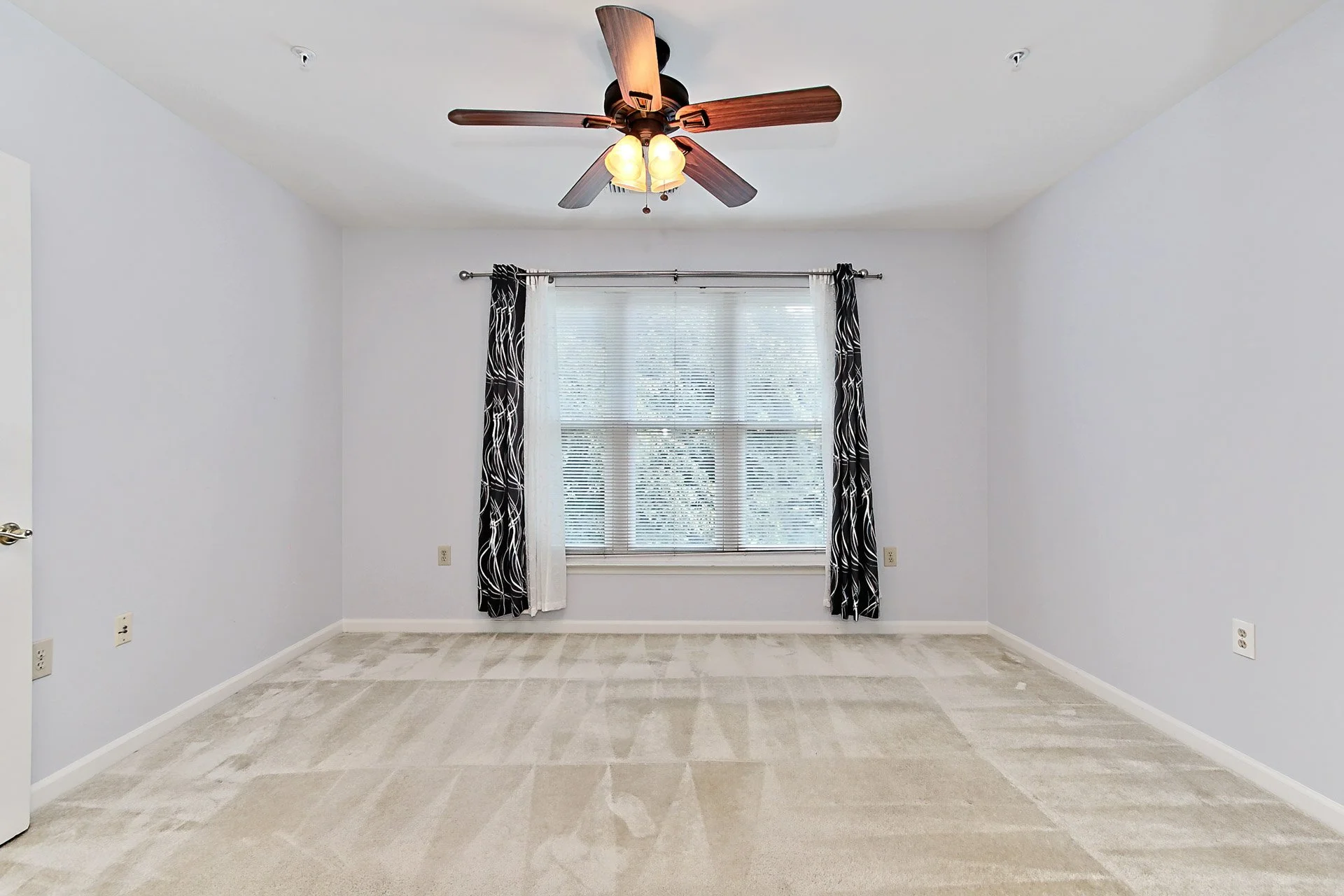 Empty bedroom with a ceiling fan, window with blinds and curtains, and beige carpet.