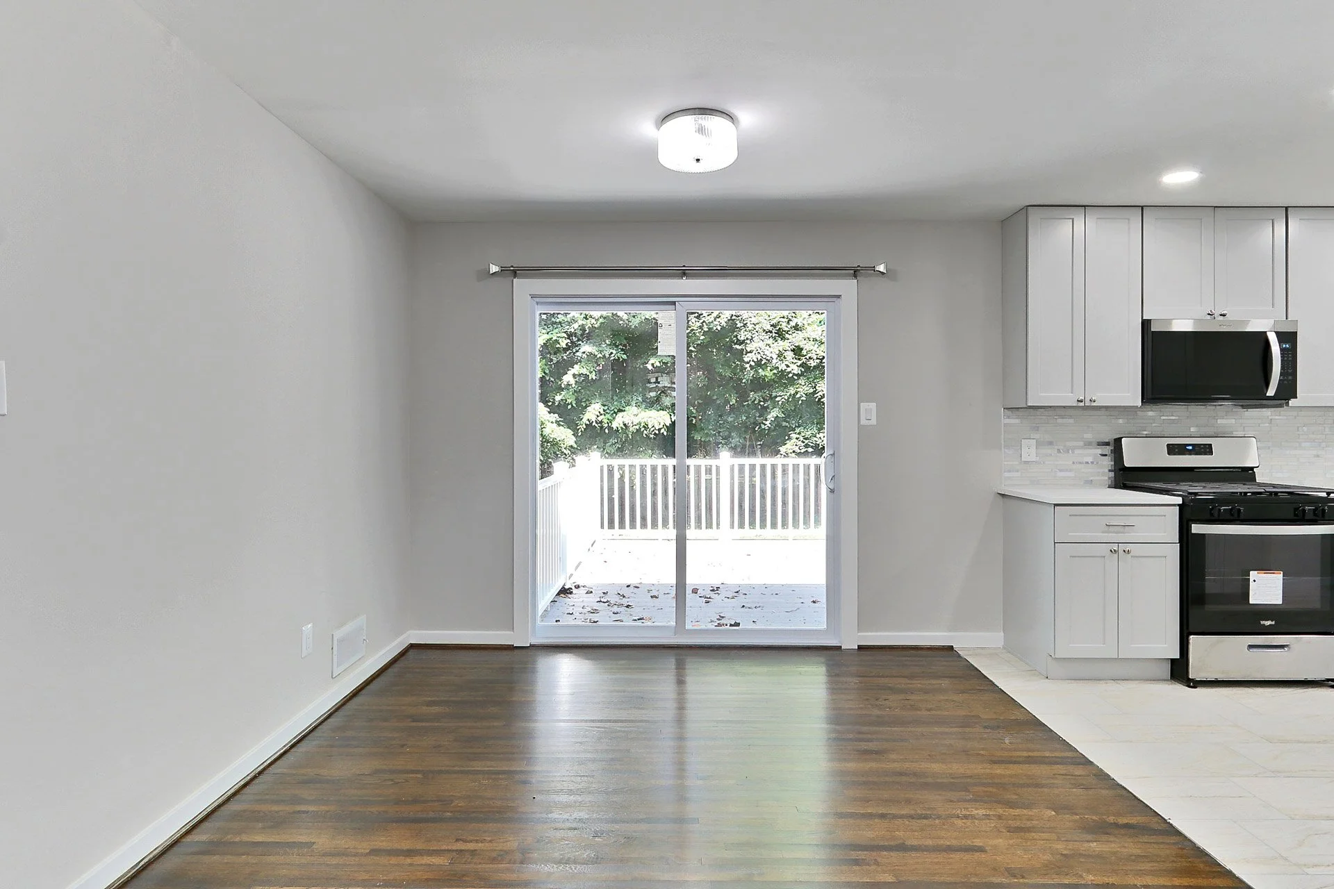 Interior view of a kitchen with a sliding glass door opening to a deck and greenery outside. The kitchen has white cabinets, a stainless steel microwave, a stove, and a backsplash. The room has two ceiling lights, and the floor is a mix of hardwood a