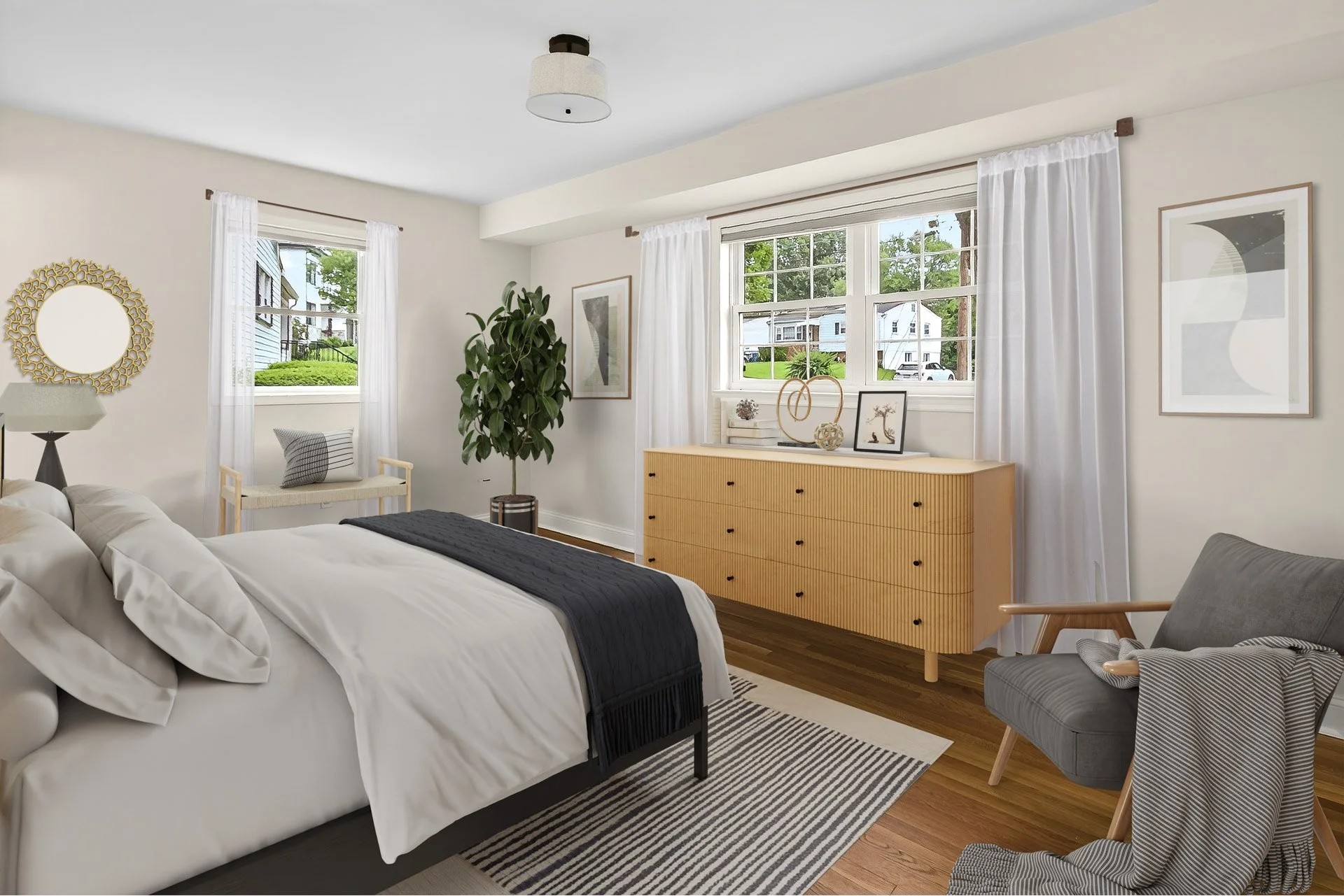 Bright bedroom with white walls, large windows with white curtains, a bed with white bedding, a black throw, and pillows, a gray armchair with a blanket, a wooden dresser with decorative items, a large plant, and framed artwork on the walls.