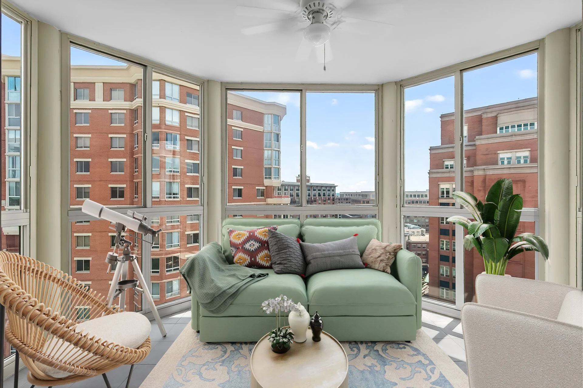 Living room with large windows showing city buildings, a green sofa with pillows, a telescope, a wicker chair, a small round table with decorative items, and a large potted plant.