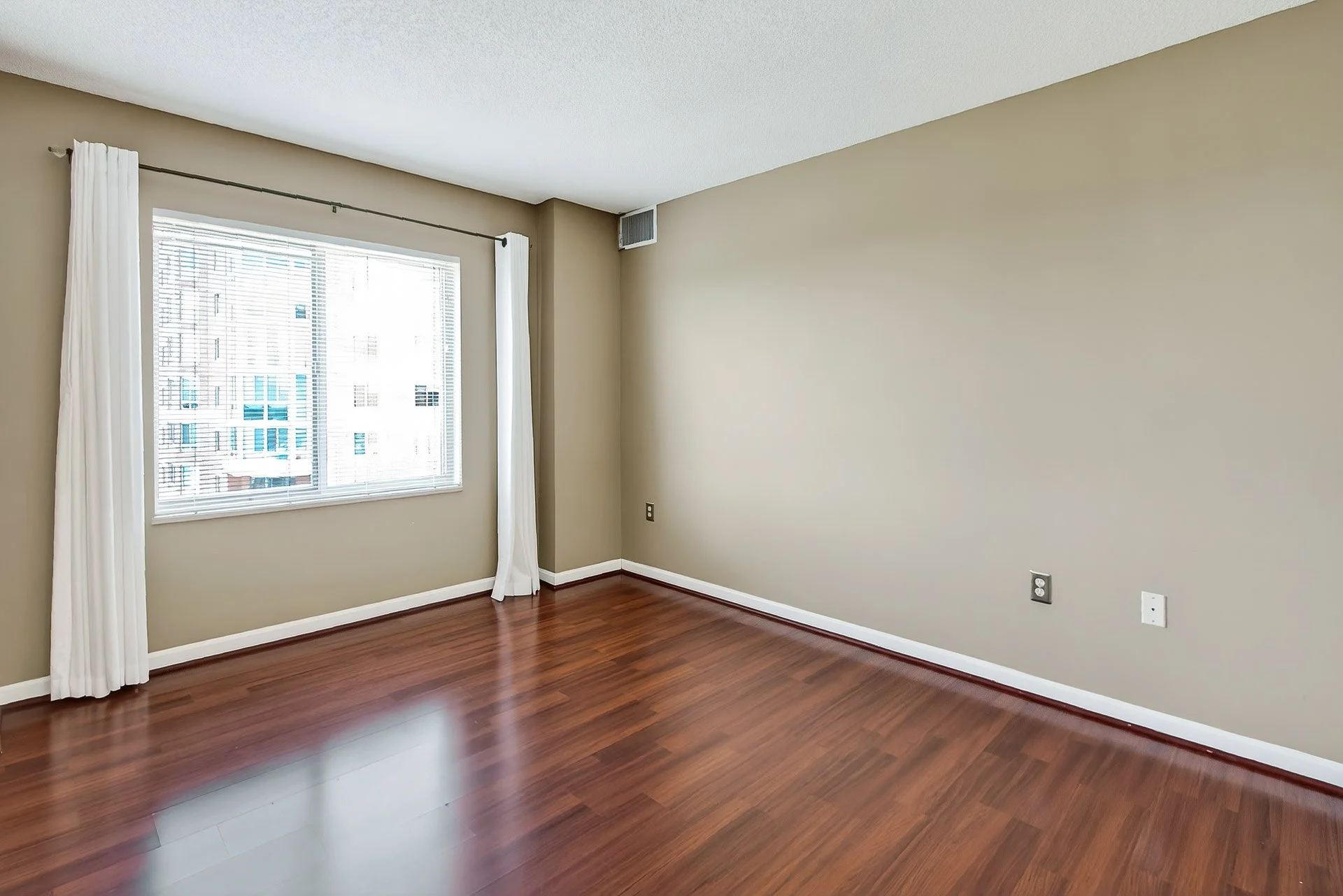 Empty room with hardwood floors, beige walls, a window with blinds and white curtains, and an air vent near the ceiling.