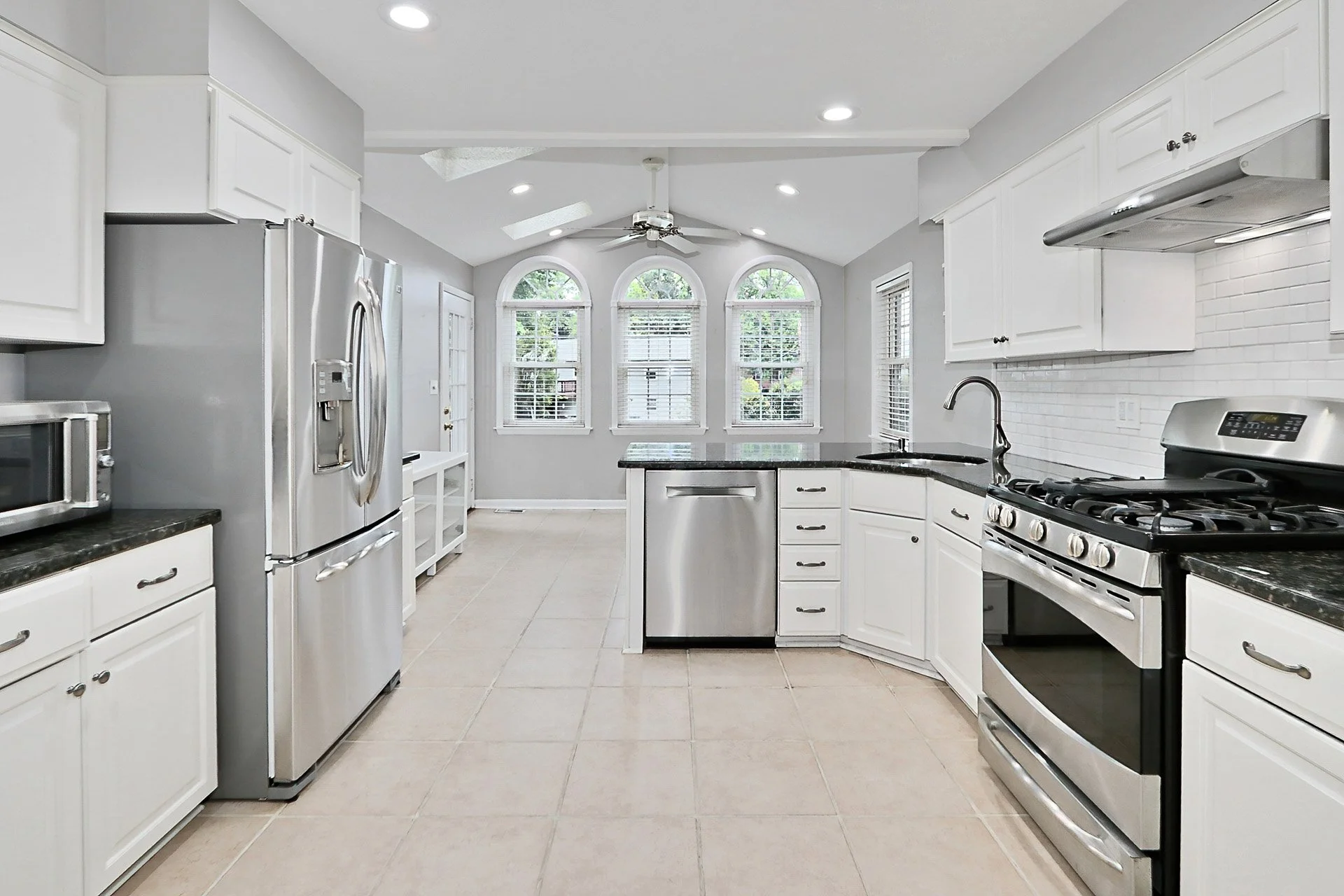 Bright kitchen with white cabinets, black countertops, stainless steel appliances, a ceiling fan, and large arched windows.