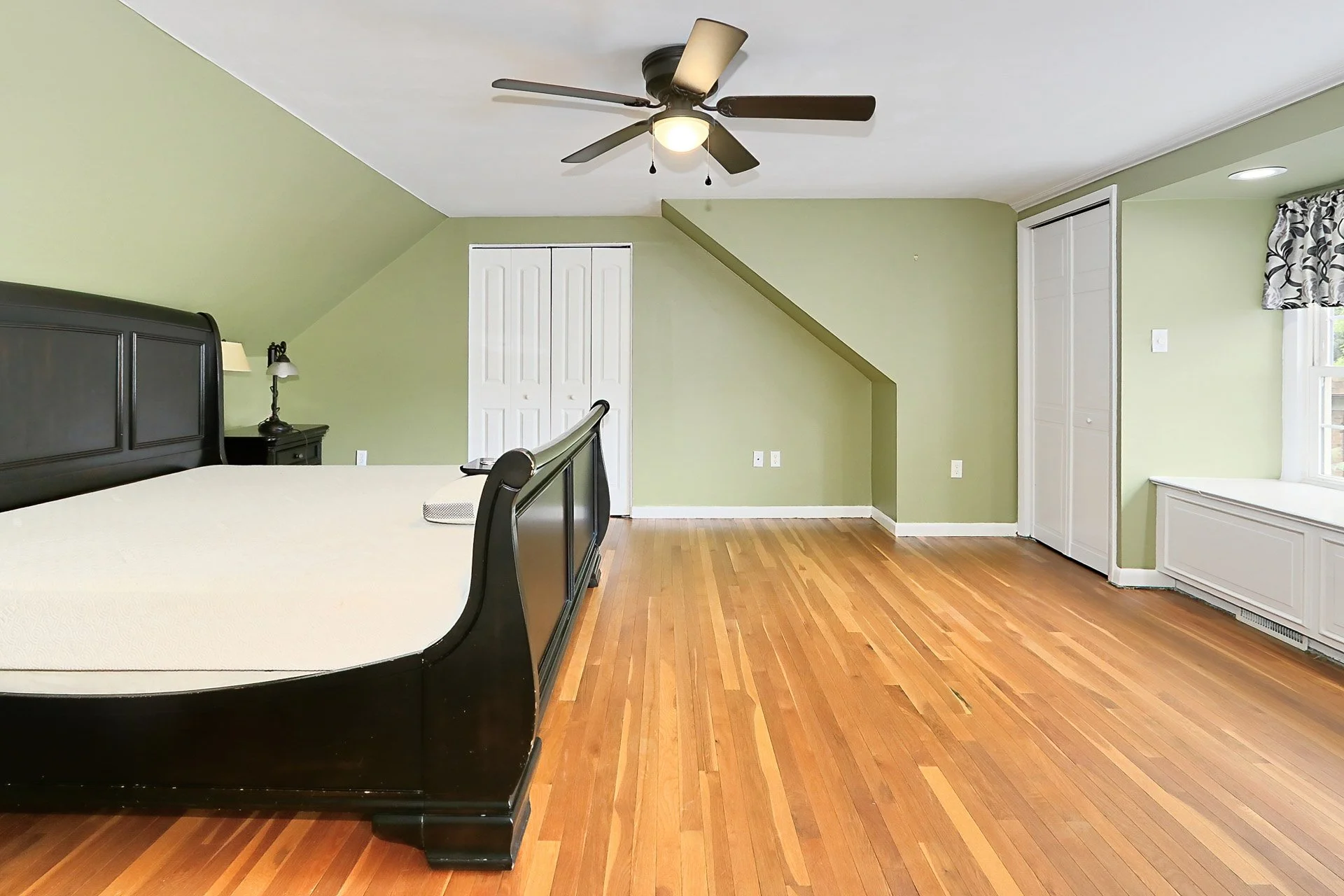 Empty bedroom with a large bed frame, hardwood floor, light green walls, ceiling fan, and window with curtains.