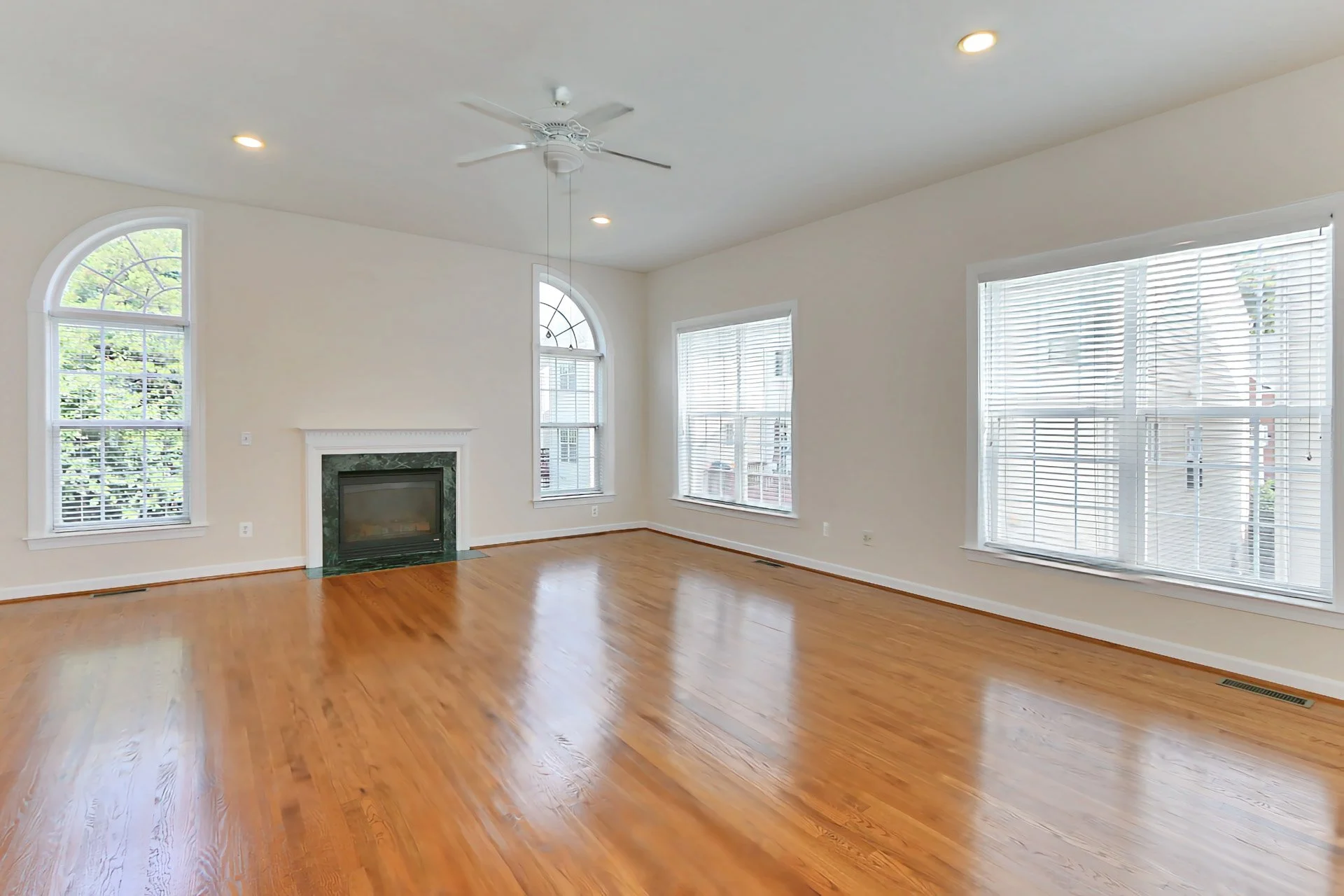 Empty living room with hardwood floors, white walls, large windows with blinds, a fireplace, a ceiling fan, and recessed lighting.