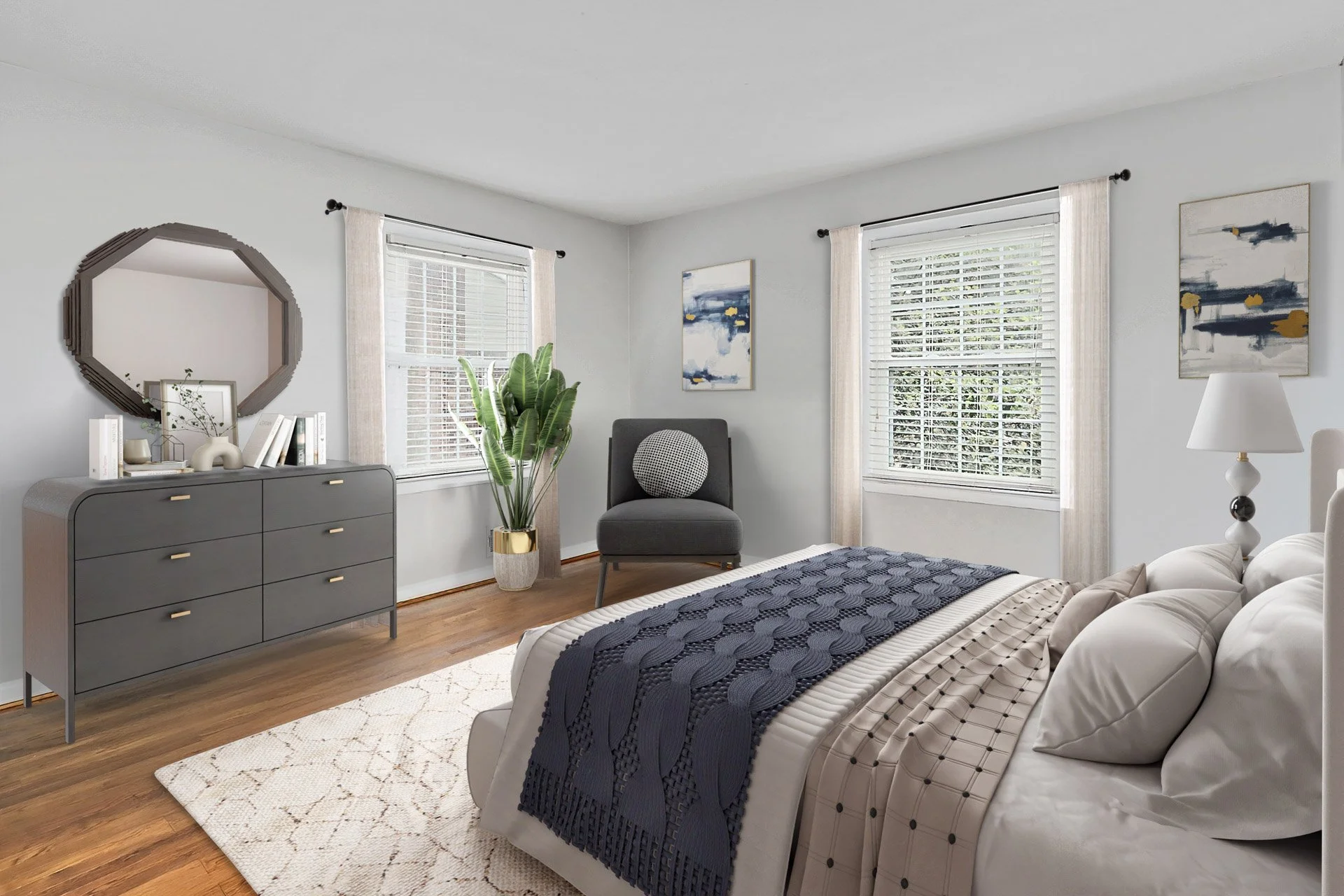 A bright bedroom with two large windows with white blinds and curtains, a white bed with a navy blue throw blanket, beige pillows, a nightstand with a lamp, a gray dresser with books and decor, a large mirror, a gray chair with a round pillow, a tall