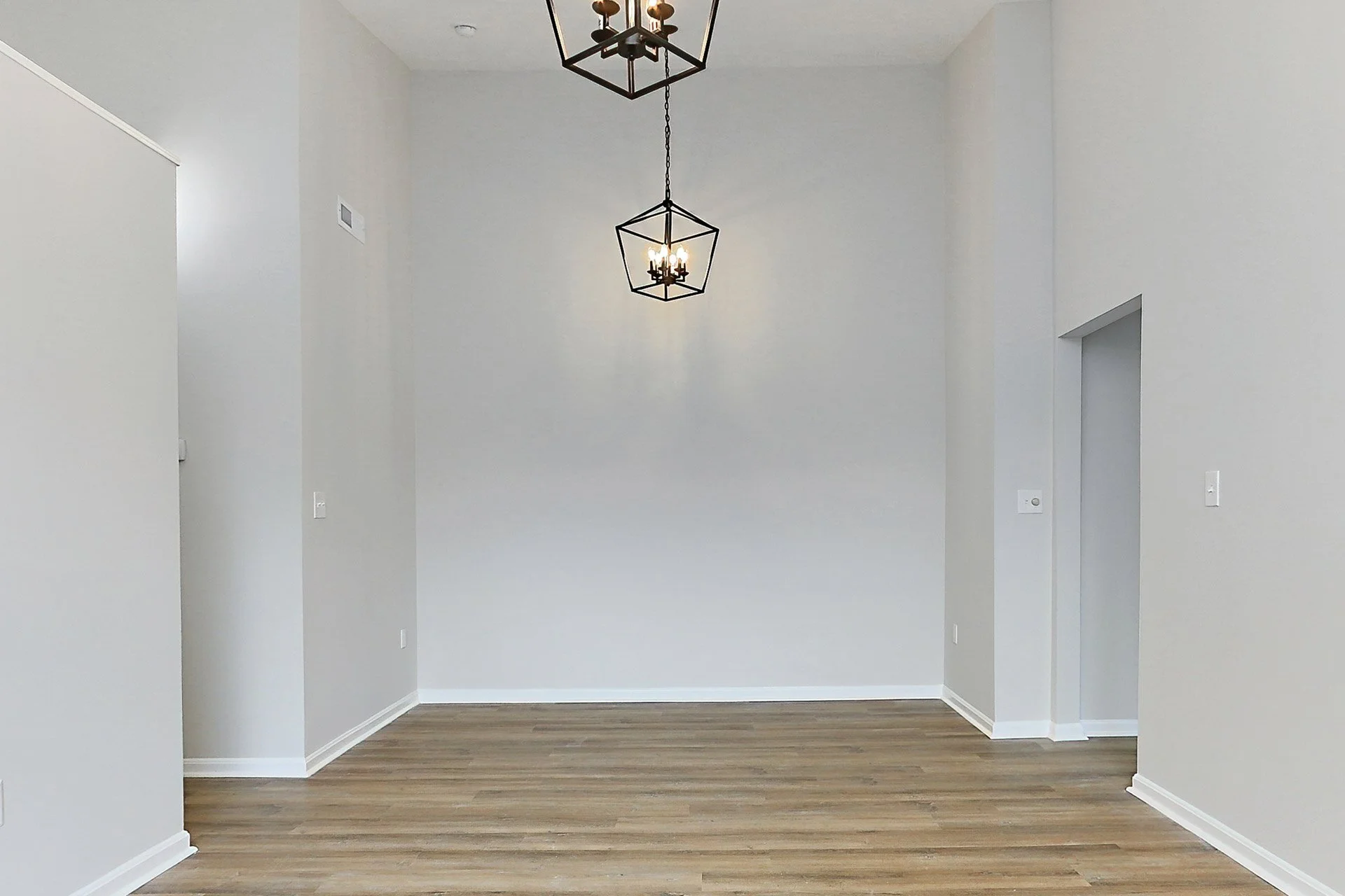 Empty room with white walls, wooden floor, and two black geometric chandeliers hanging from the ceiling.