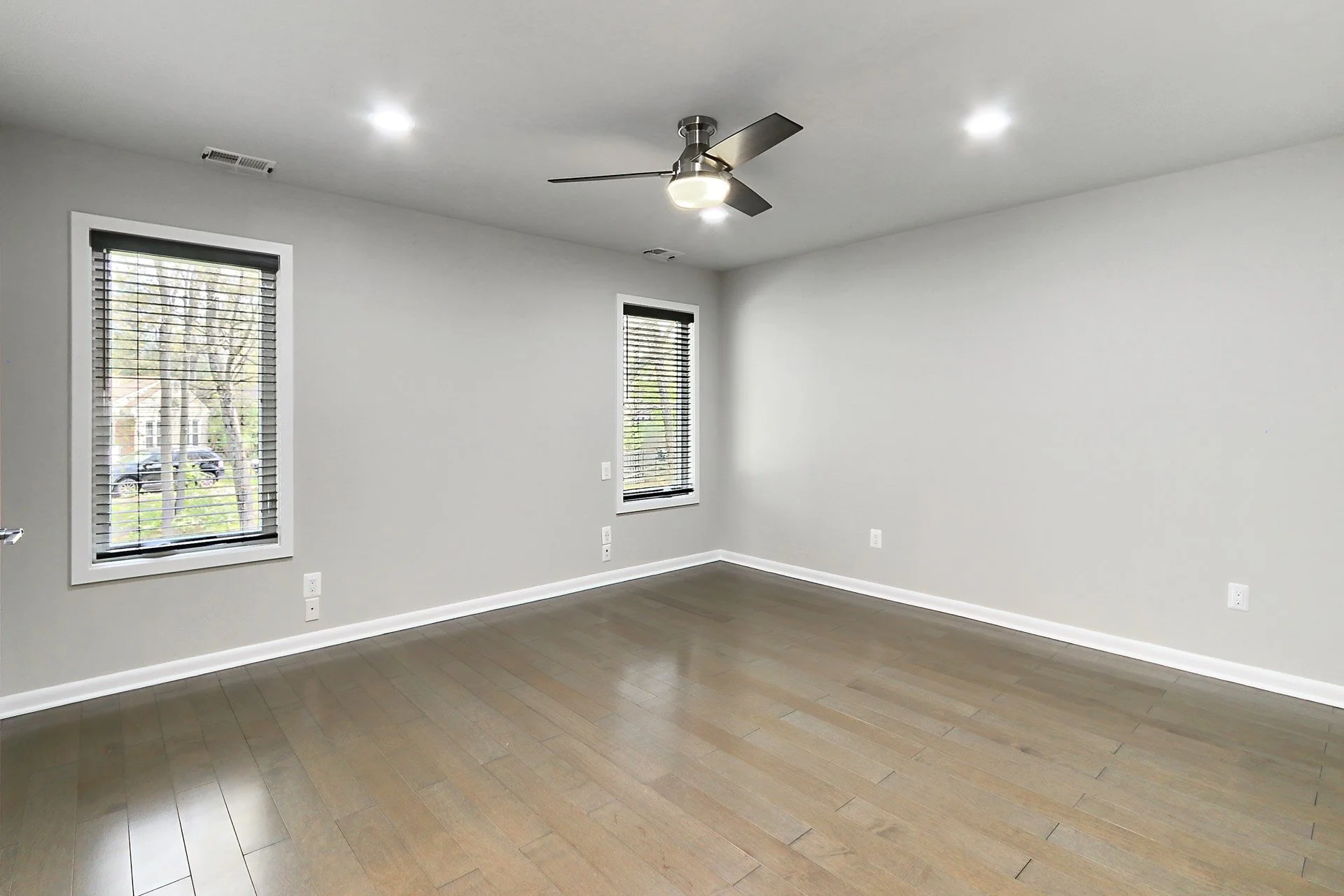Empty room with light gray walls, wooden flooring, two windows with black blinds, a ceiling fan with light, and white baseboards.