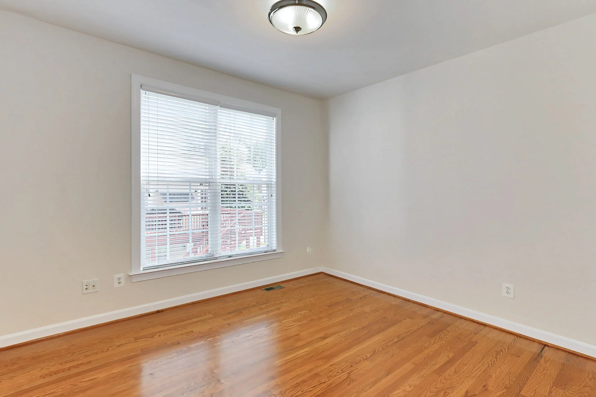 Empty room with white walls, hardwood floor, a window with blinds, and a ceiling light fixture.