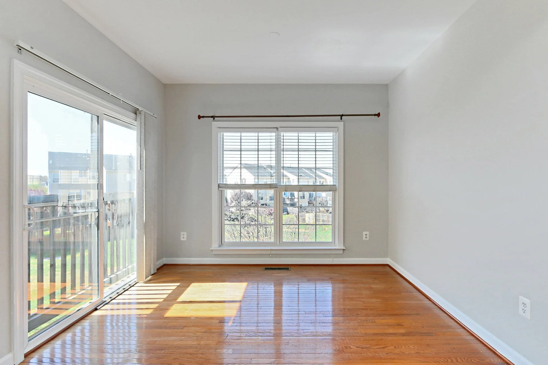 Empty living room with hardwood floors, large window with blinds, sliding glass door leading to balcony, and neutral wall colors.