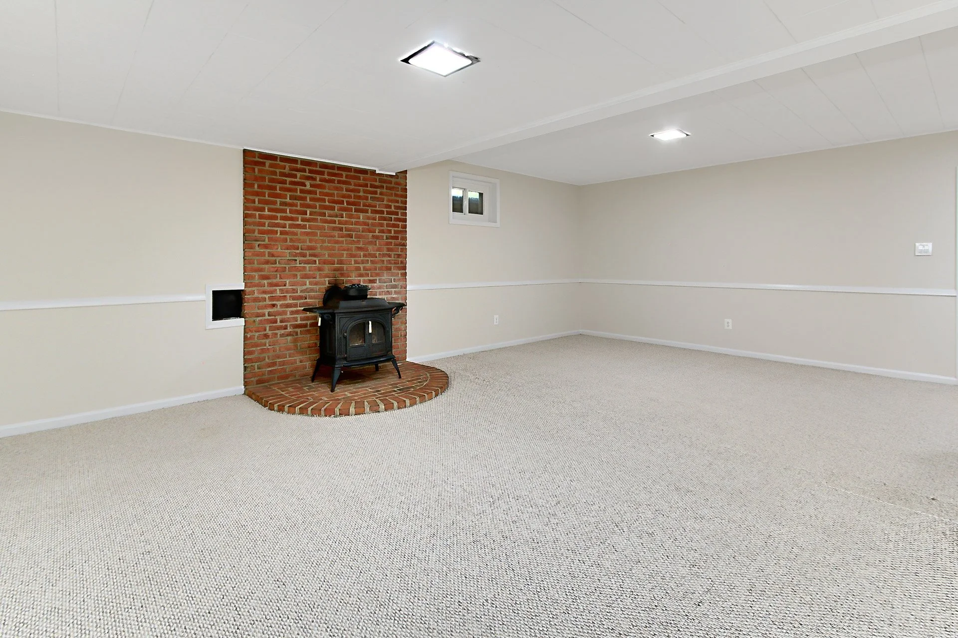Empty living room with beige carpet, white walls, a small window, ceiling lights, and a brick fireplace with a black wood stove.
