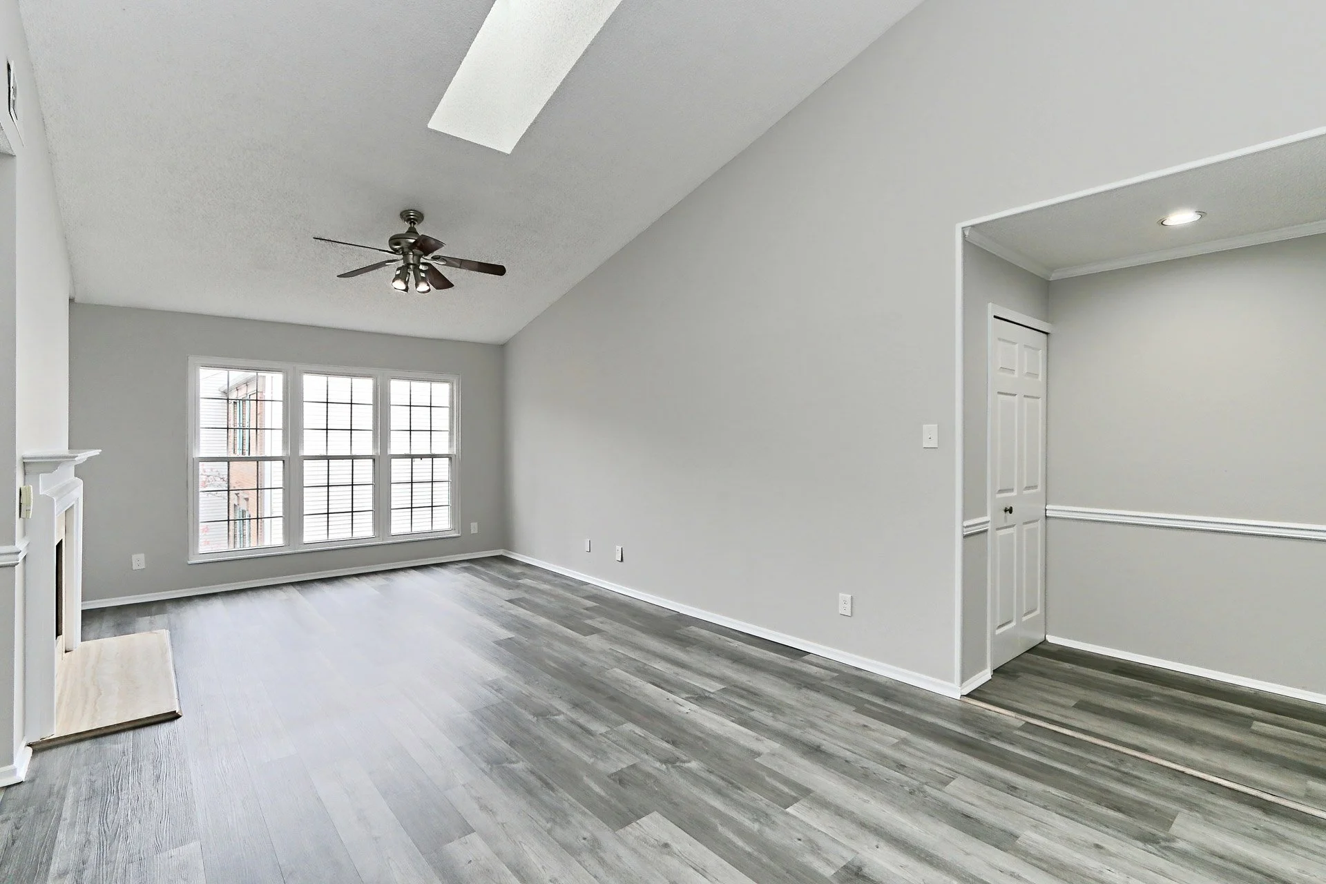 Empty living room with large front window, grey wood flooring, white walls, ceiling fan, and a small fireplace.