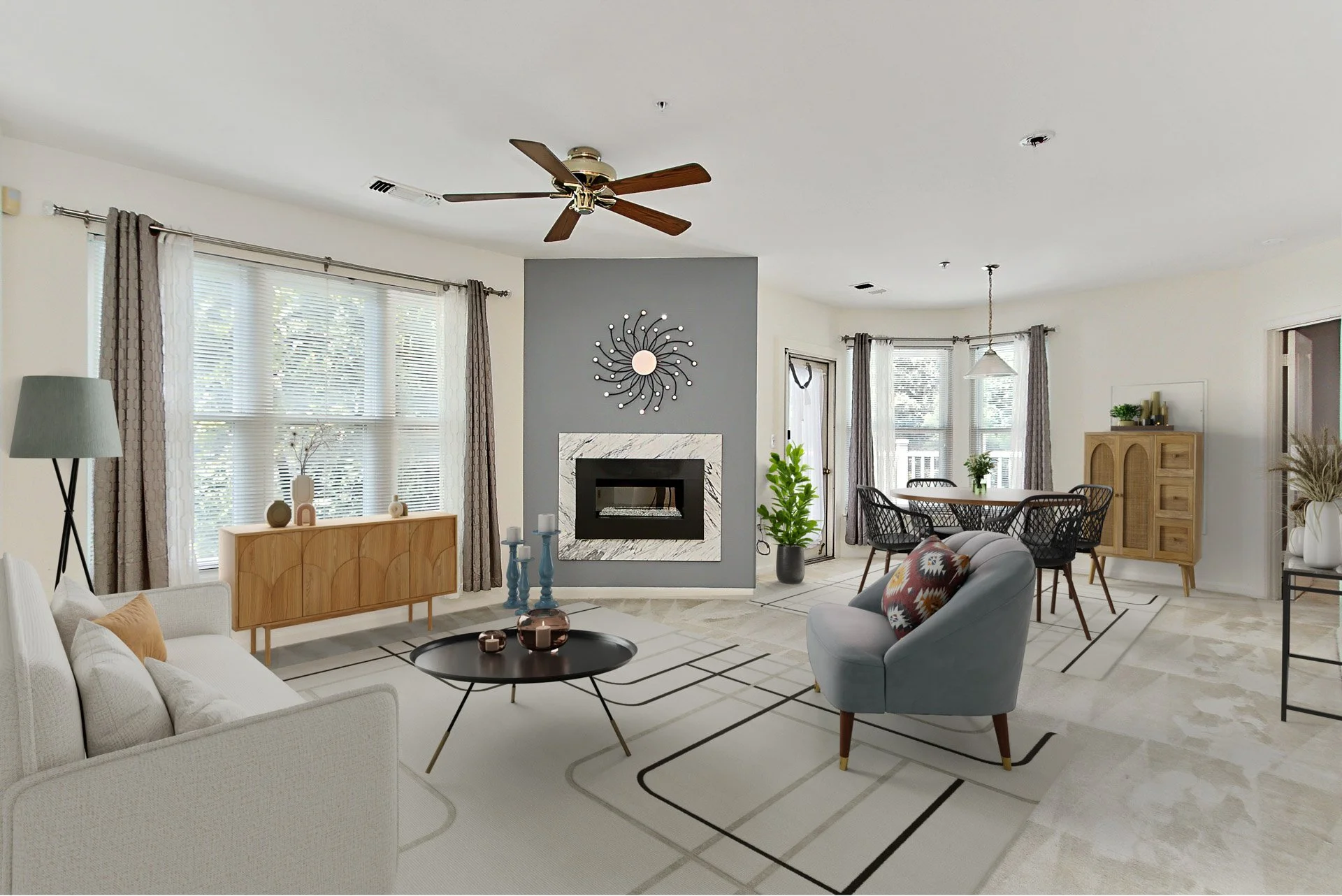 Living room with a ceiling fan, fireplace with decorative wall art, beige sofa, gray armchair with a colorful pillow, round black coffee table, dining area with black chairs, and large windows with curtains.
