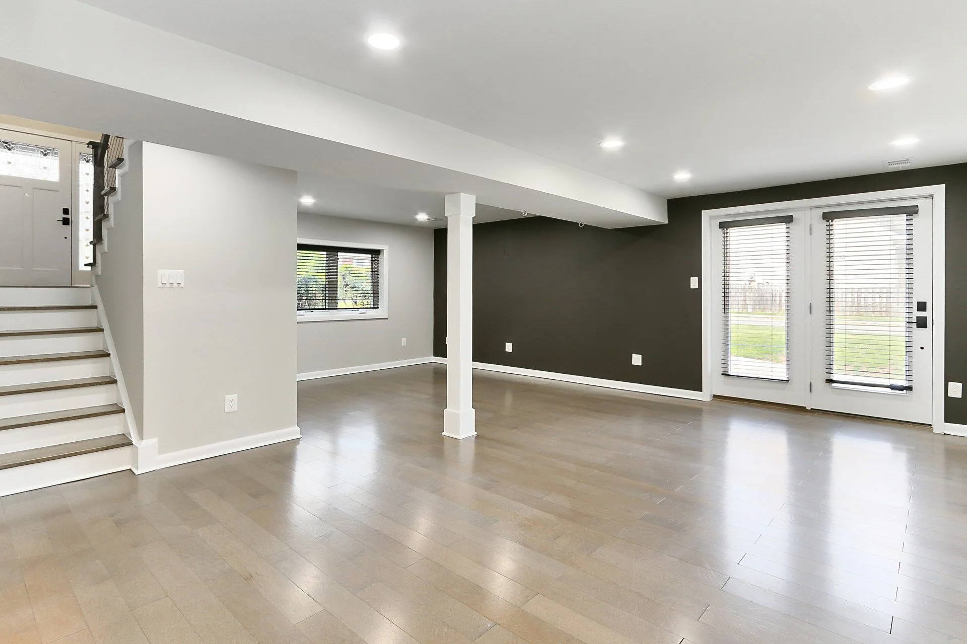 Empty living room with light-colored hardwood floors, white and dark gray walls, a staircase in the corner, and multiple windows and glass doors with blinds.