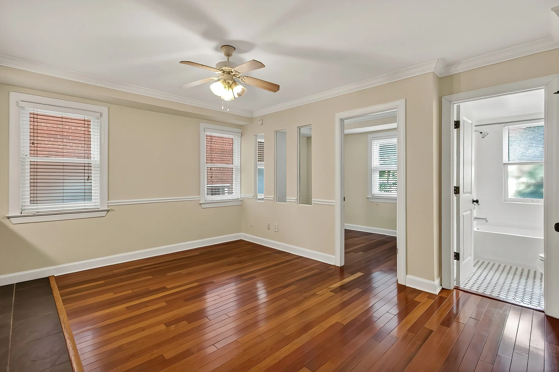 Empty room with hardwood flooring, three windows with blinds, ceiling fan with lights, doorway leading to a bathroom with a bathtub and a window.