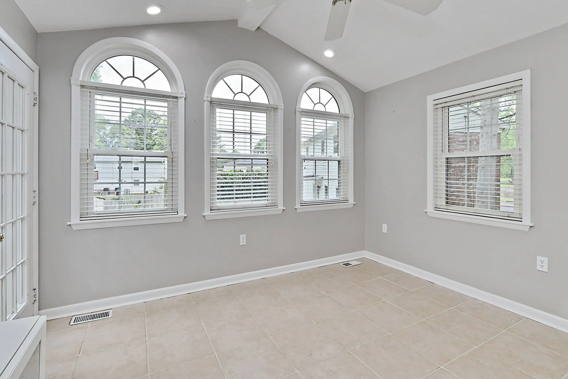 Empty room with four large arched windows with white blinds, light gray walls, beige tiled floor, and a ceiling fan.