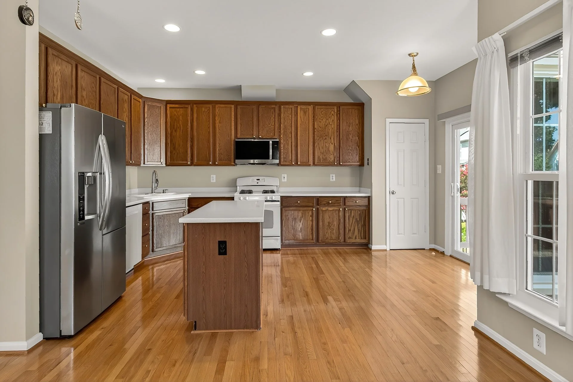 Empty kitchen with wooden cabinets, stainless steel refrigerator, white countertops, and hardwood floors. There are windows with white curtains and a door leading outside.