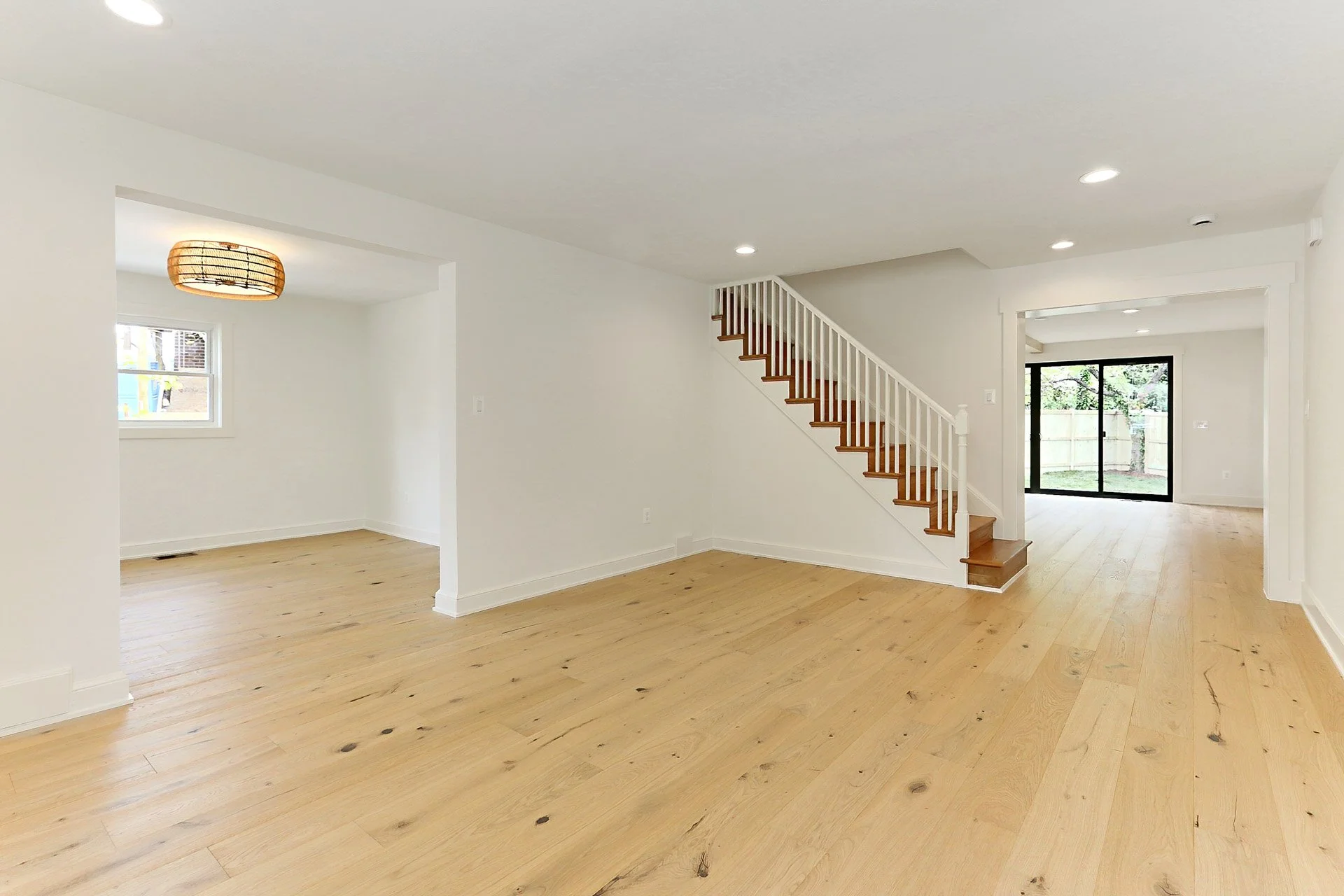 Empty living room with light wooden floors, white walls, a staircase with wooden steps and white railing, and large sliding glass doors leading outside.
