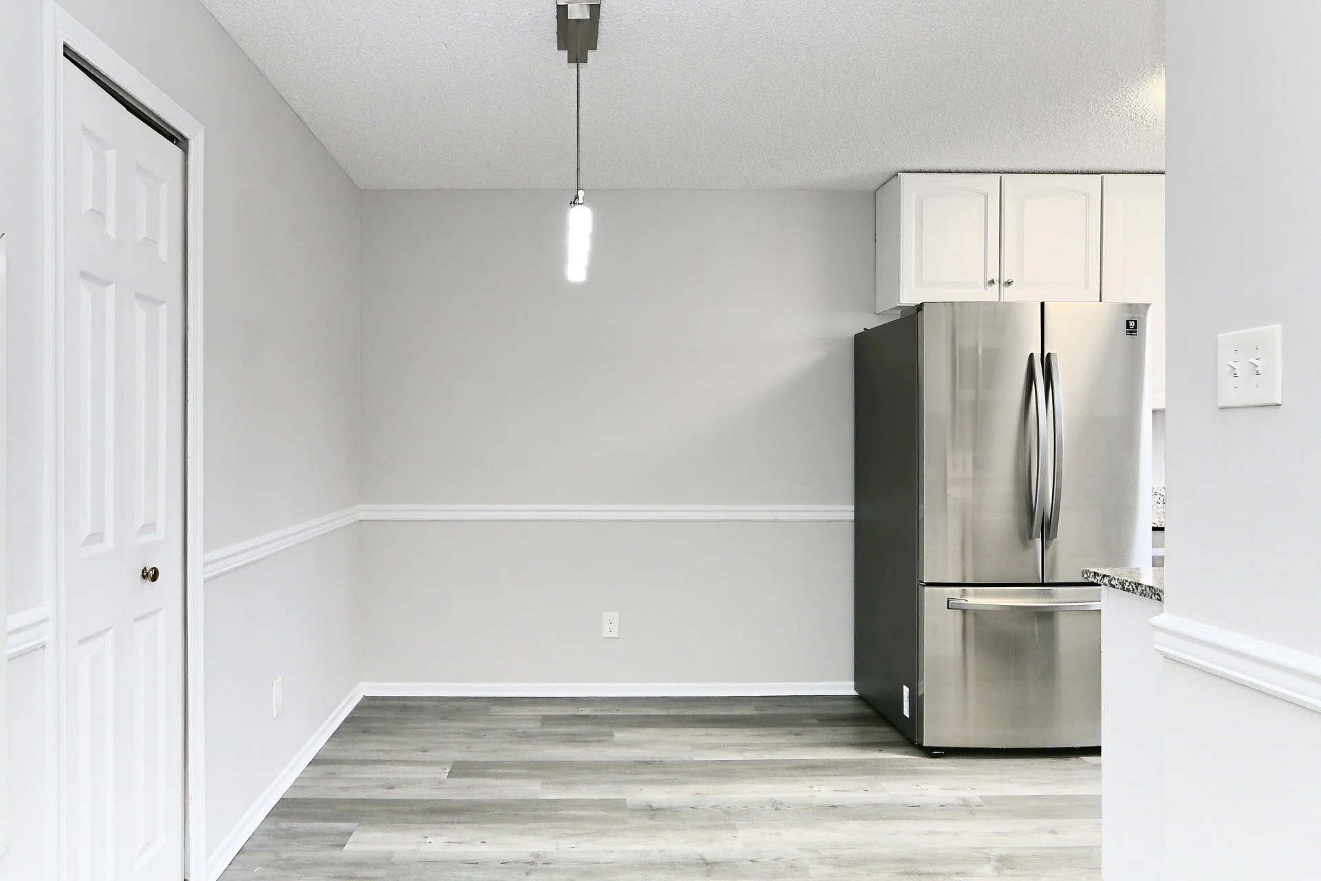 Empty kitchen corner with gray walls, gray wood flooring, a white ceiling, a small hanging light, and a stainless steel refrigerator with white cabinets above.