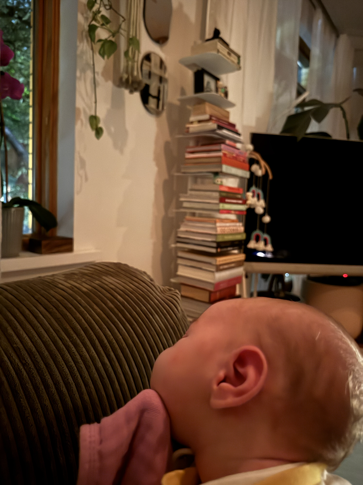 A close-up of a baby sleeping on a striped brown and beige couch with a bookshelf, a TV, and hanging decorations in the background.