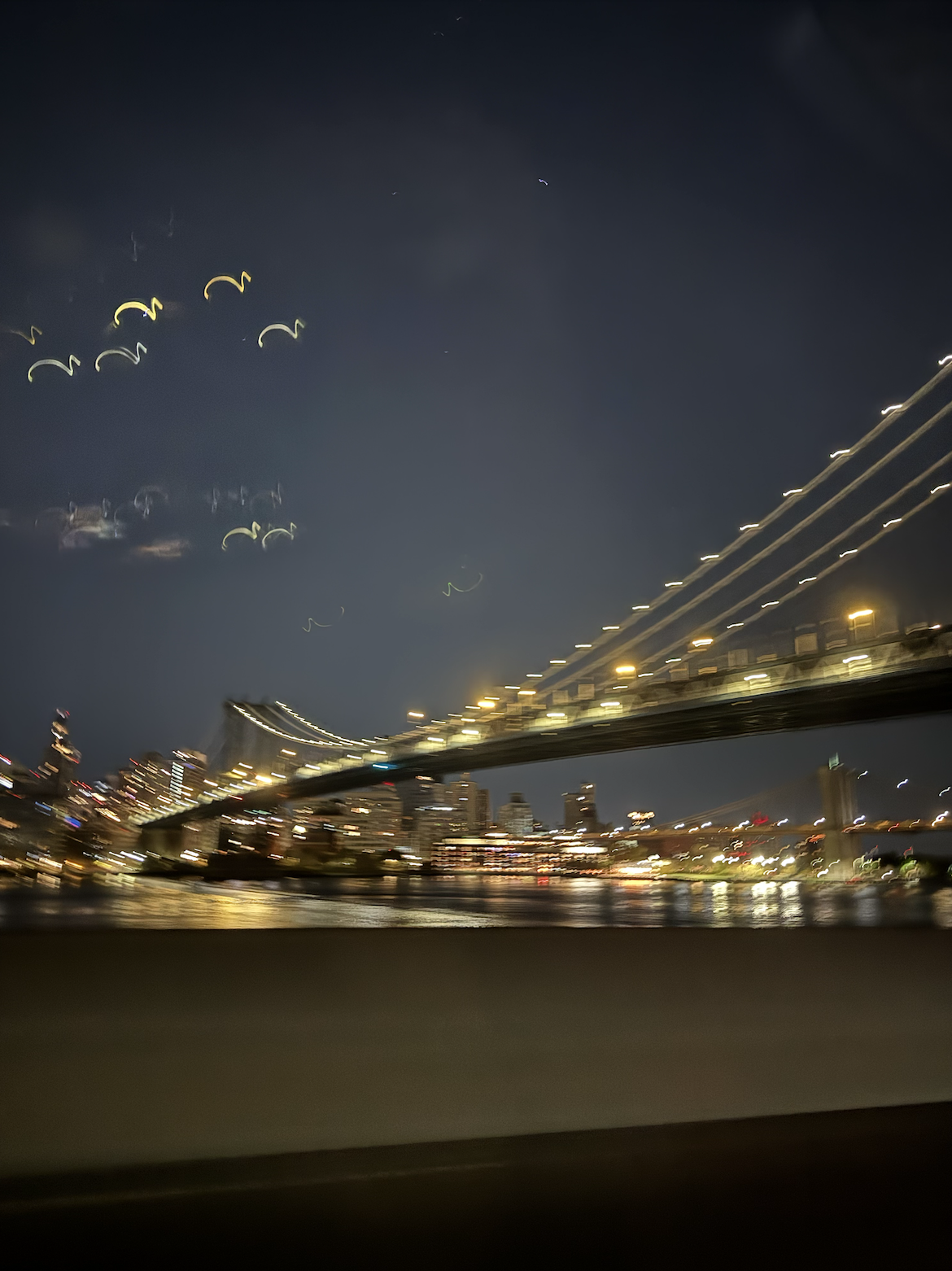Nighttime cityscape with illuminated bridge over water and blurred city lights in background.