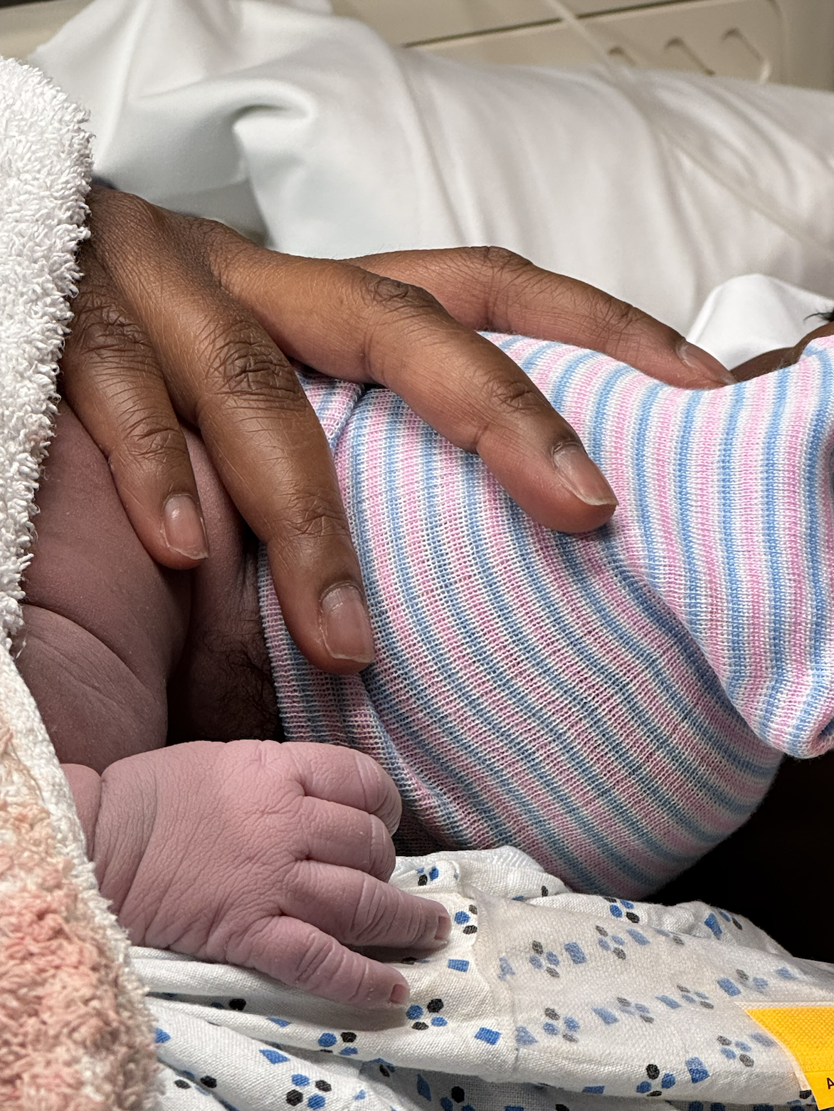 Close-up of a newborn baby and an adult's hand gently touching, with the baby wearing a striped pink, blue, and white outfit, lying on hospital bedding.