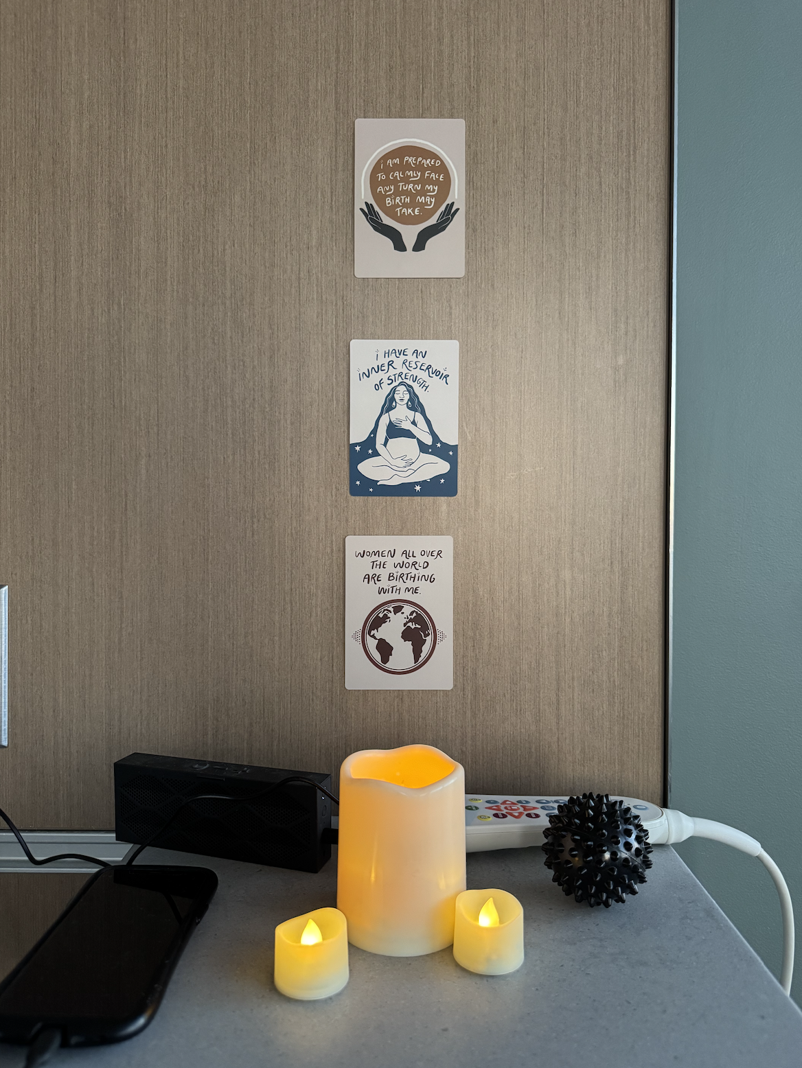 Decorative candles and a remote control on a table in front of a wall with three inspirational posters about strength and womanhood.
