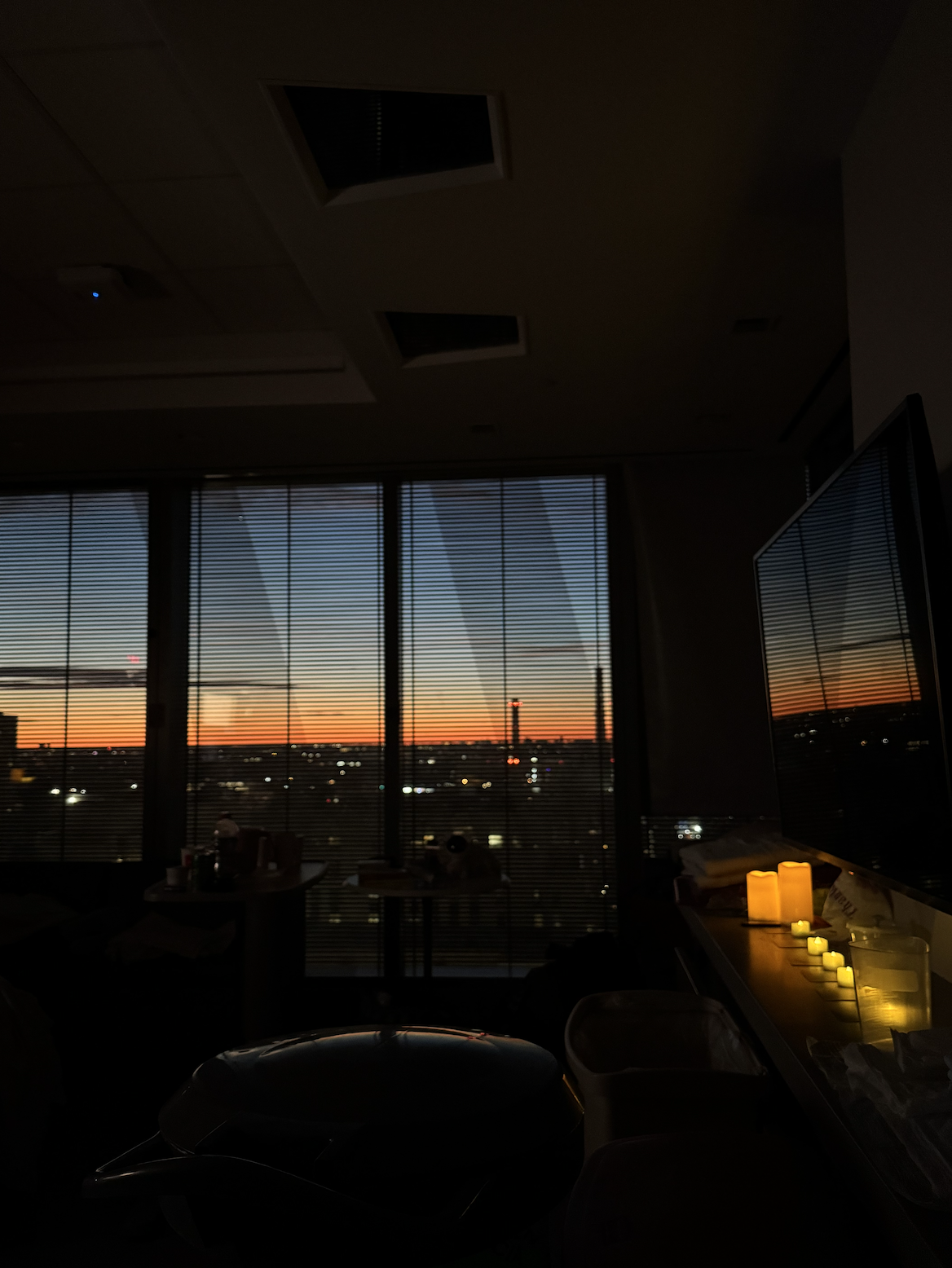 Room with large window blinds showing a sunset with orange and blue sky, and a cityscape with buildings and lights outside. Inside, there are lit candles on a table or counter along with various objects and equipment.