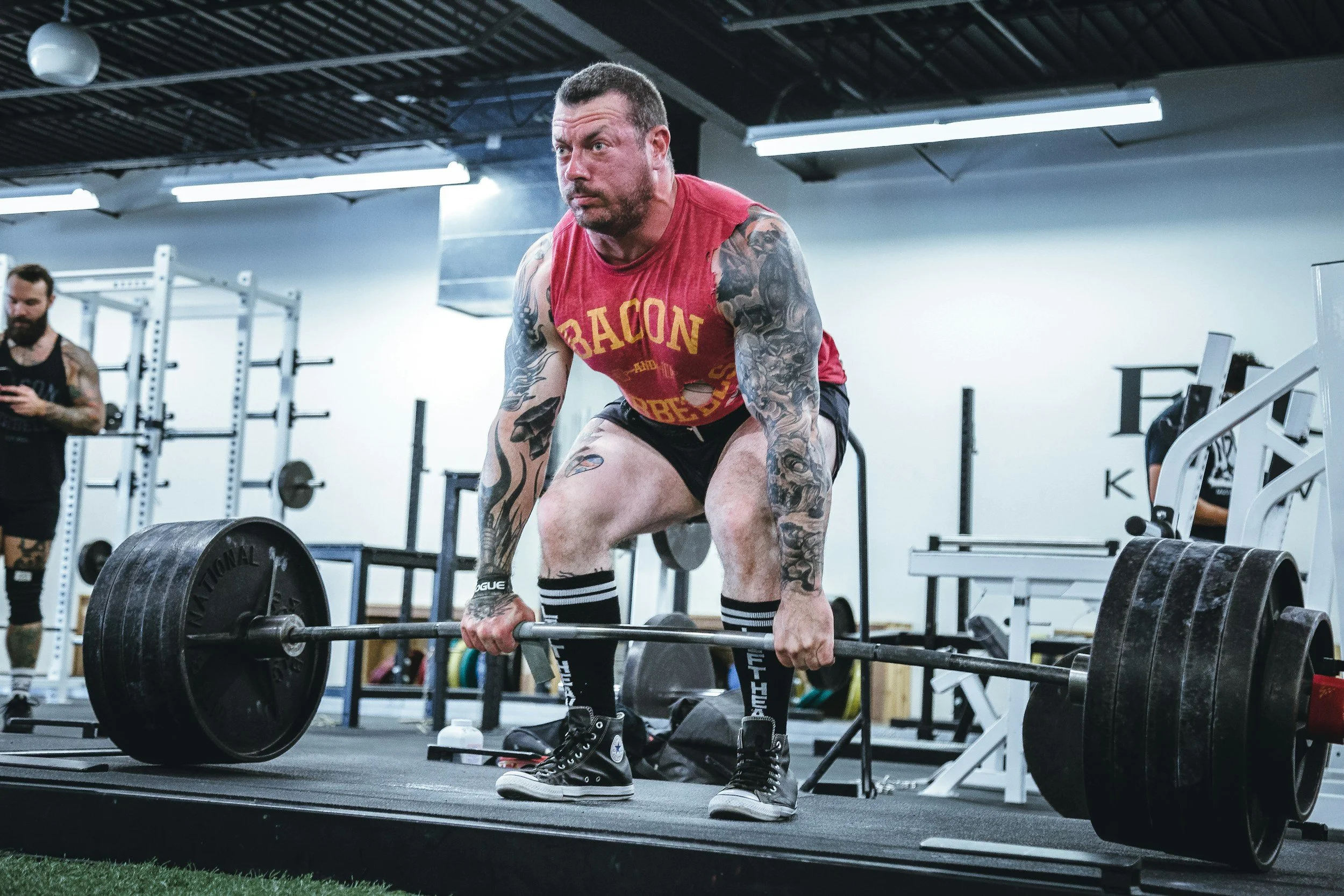 A muscular man with tattoos lifting a heavy barbell in a gym, wearing a red sleeveless shirt and black shorts.
