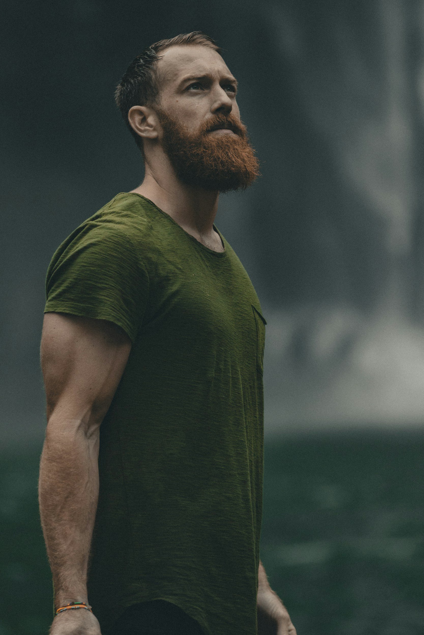 A man with a red beard and short, slicked-back hair looks to the side in a dark outdoor setting, wearing a green t-shirt.