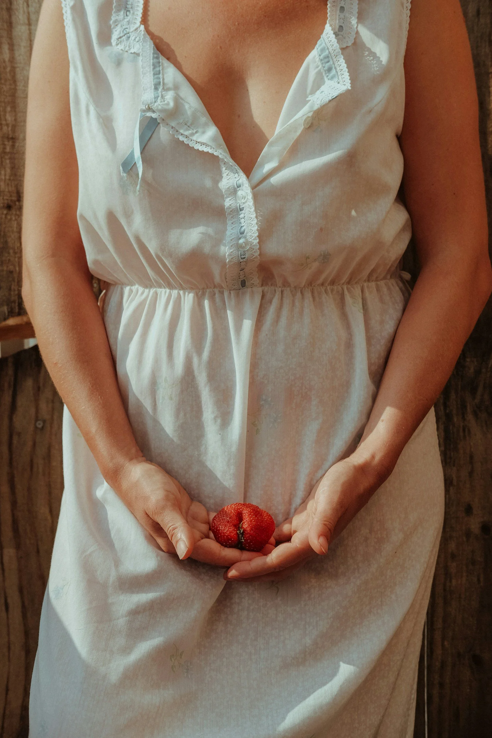 A woman in a white dress holding a red strawberry in her hands, standing against a wooden background.