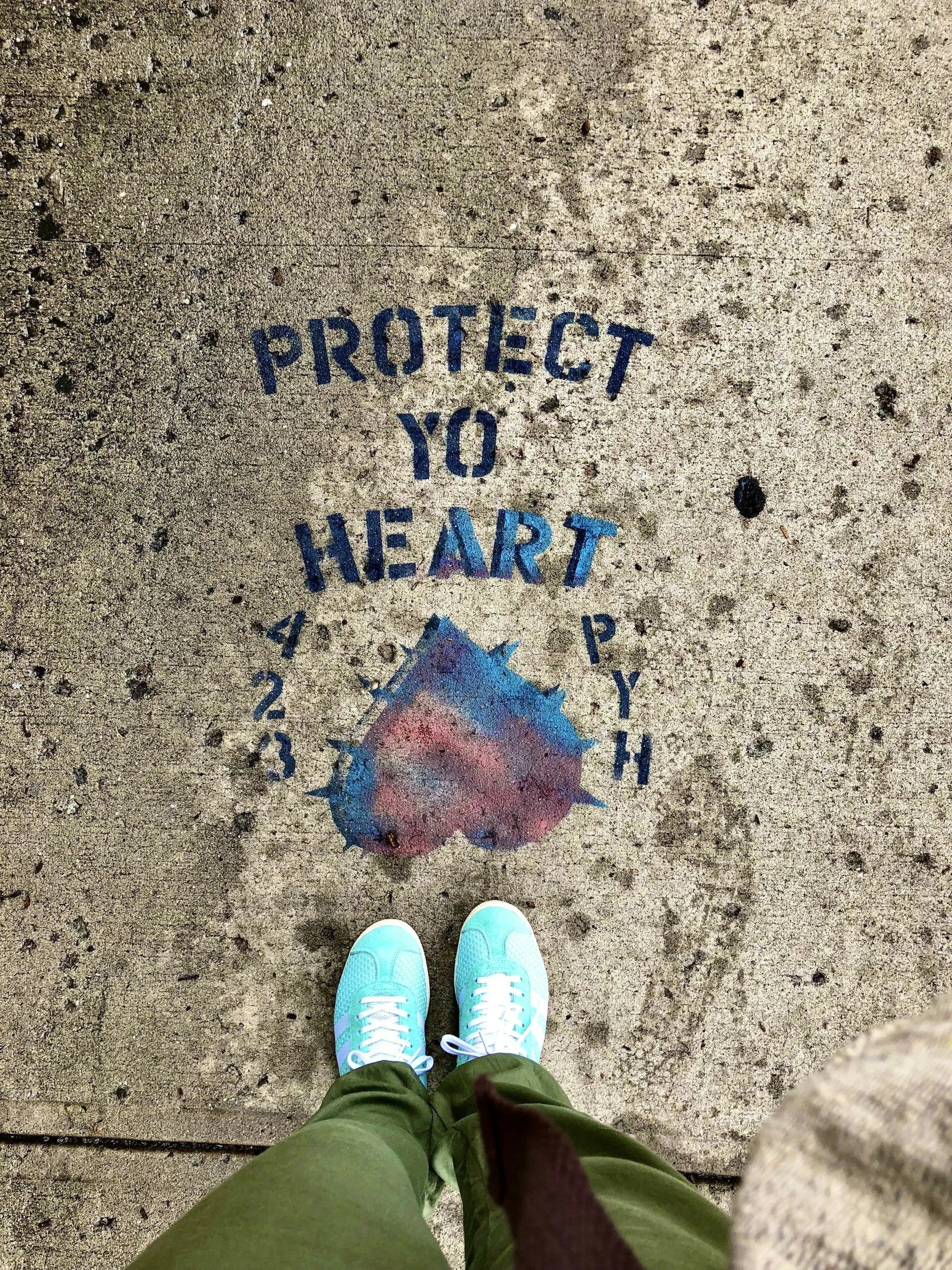 Concrete sidewalk with a painted message saying 'Protect Your Heart,' featuring a stylized heart with a spiked outline and numbers 1 to 4 around it, and a person wearing teal shoes standing in front of the message.