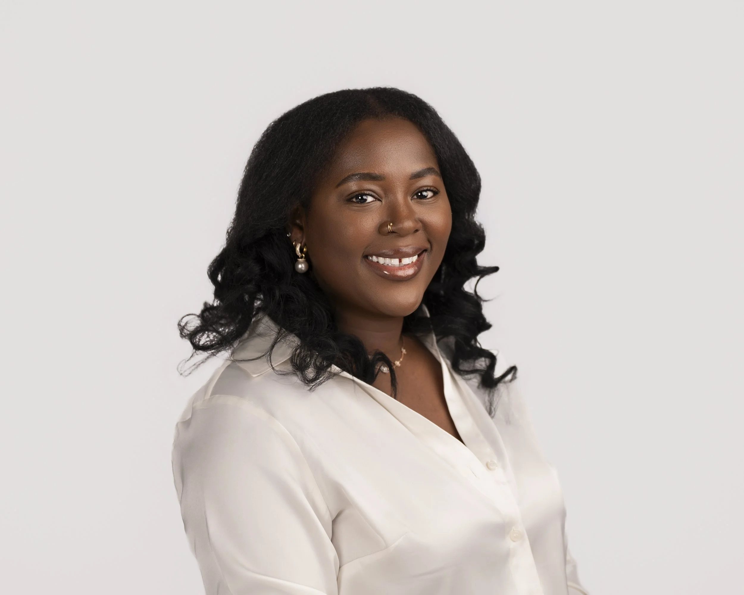 Portrait of a smiling African American woman with dark curly hair, wearing pearl earrings and a white button-up shirt, against a plain white background.