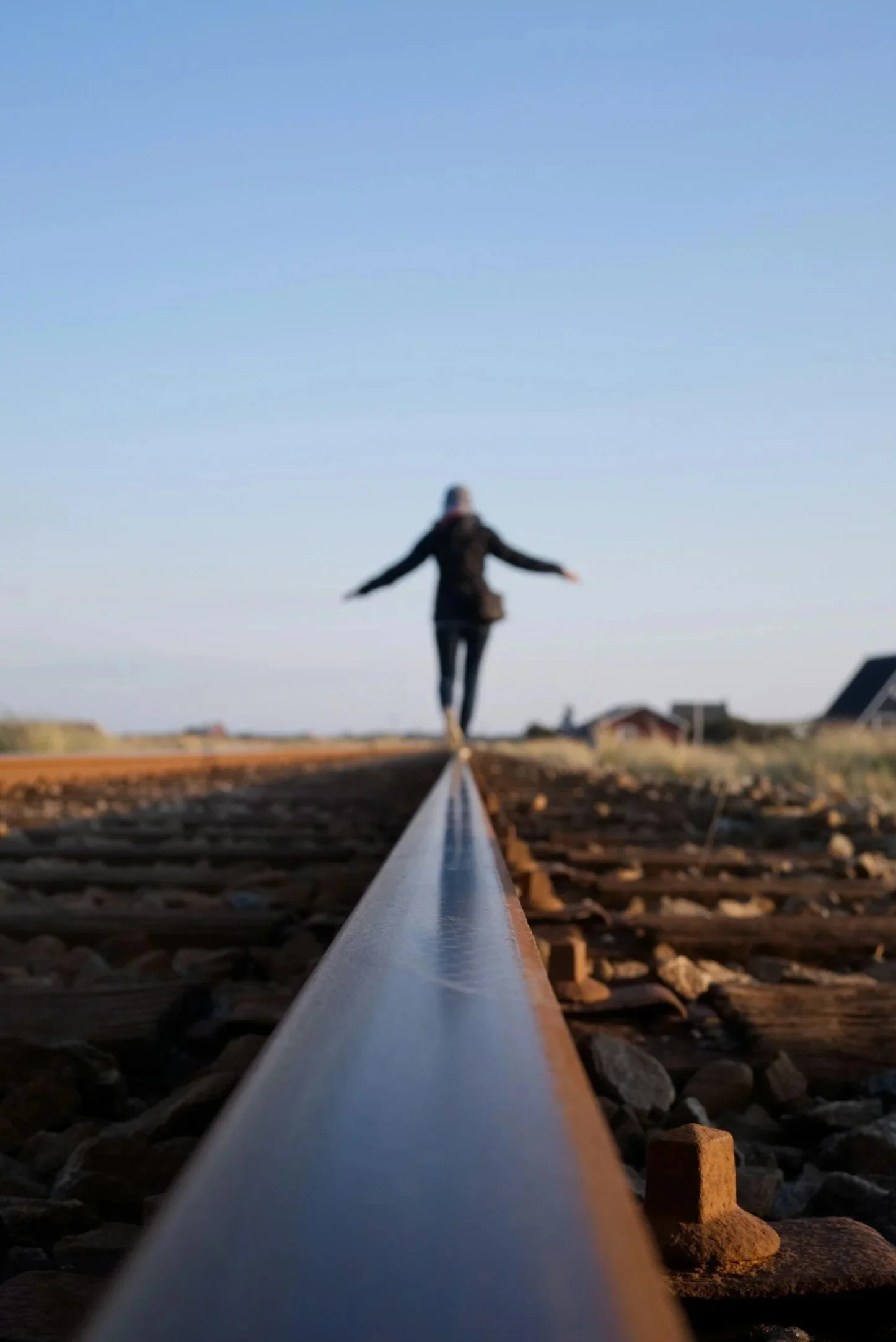 Person balancing on a railroad track outdoors during daylight, with buildings and sky in the background.