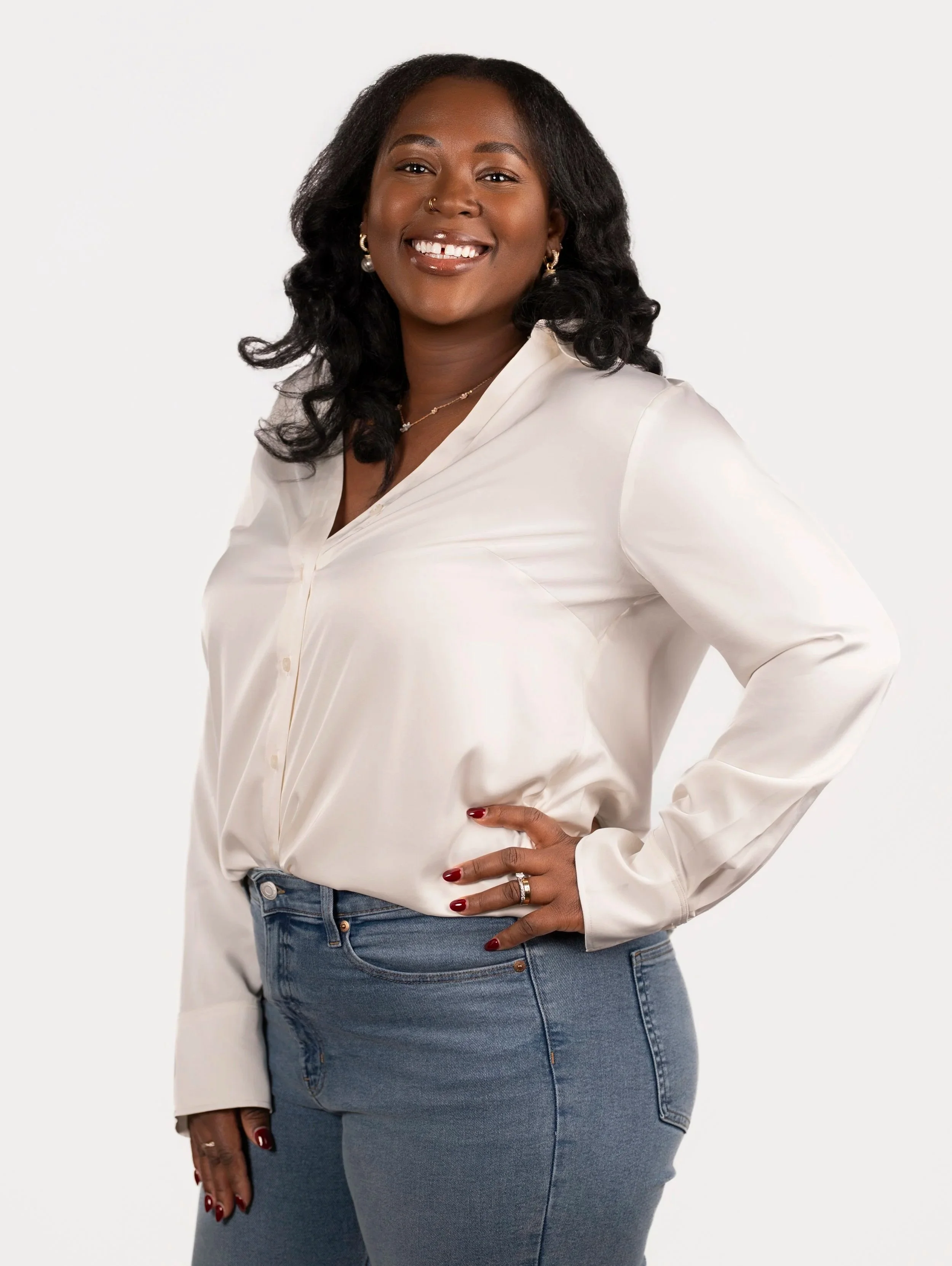 A woman with dark hair and dark skin, smiling, wearing a white blouse and jeans, with jewelry, standing against a light background.