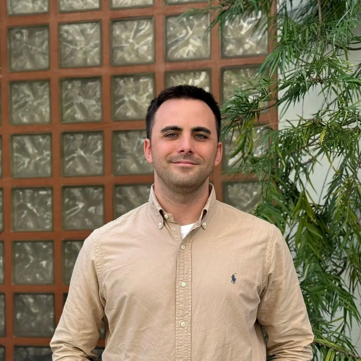 A man with short dark hair and light skin, wearing a beige button-up shirt with a small logo, standing outdoors in front of a glass block wall and a green leafy plant.