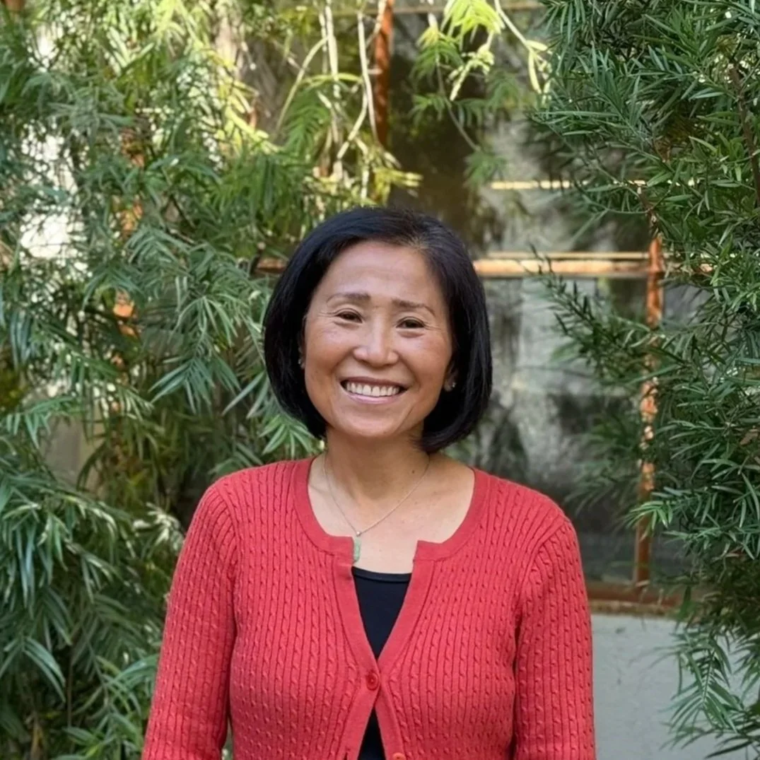 A woman with short black hair smiling in a garden with green plants, wearing a red cardigan over a black top.