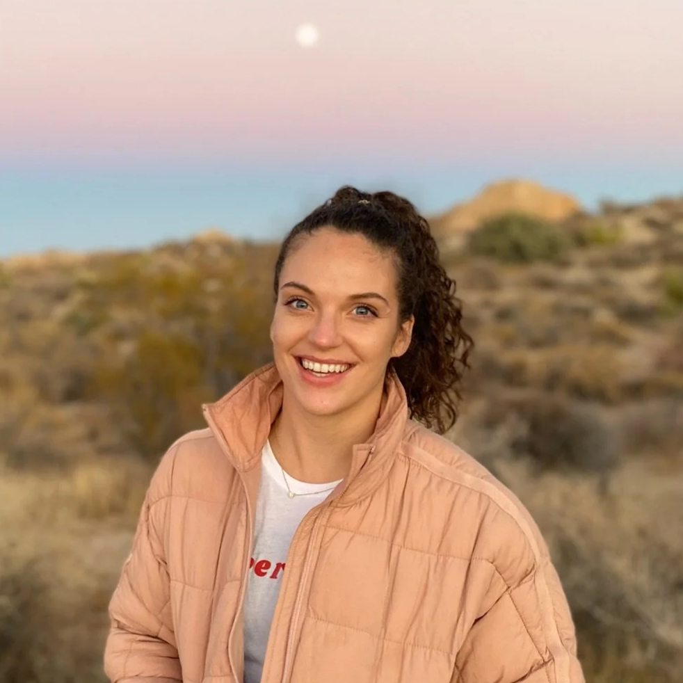 Young woman with curly brown hair smiling outdoors during sunset, wearing a peach-colored puffer jacket and a white T-shirt, with desert landscape and mountains in the background.
