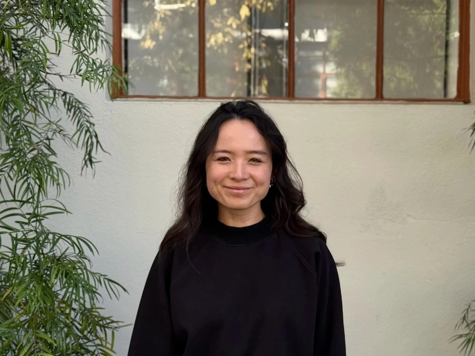 A woman with black wavy hair, smiling, wearing a black top, standing outdoors against a white wall with wooden-framed windows and greenery.
