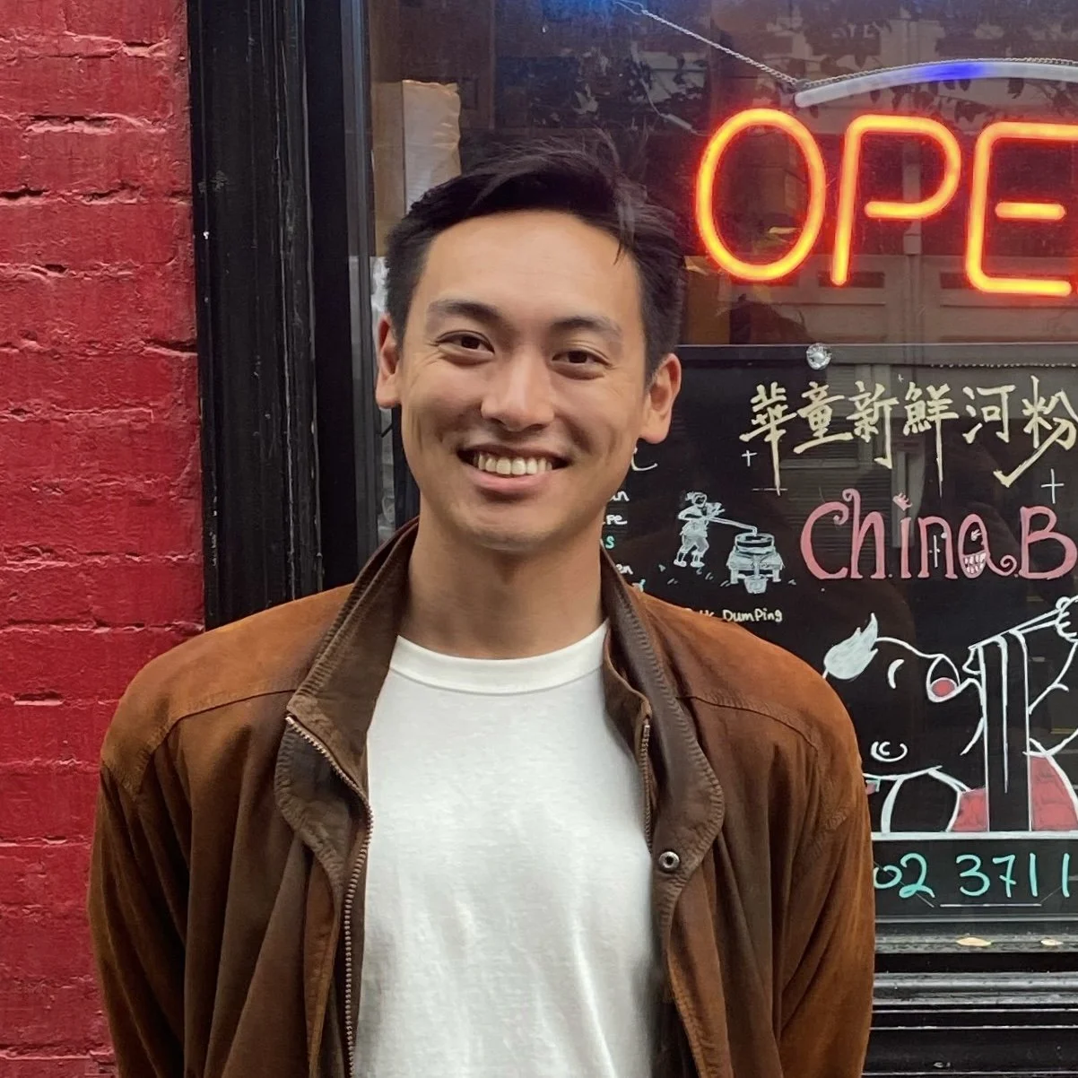 A smiling man standing outside a storefront with an illuminated "OPEN" sign and a window display that includes Chinese characters and illustrations of a person playing drums.