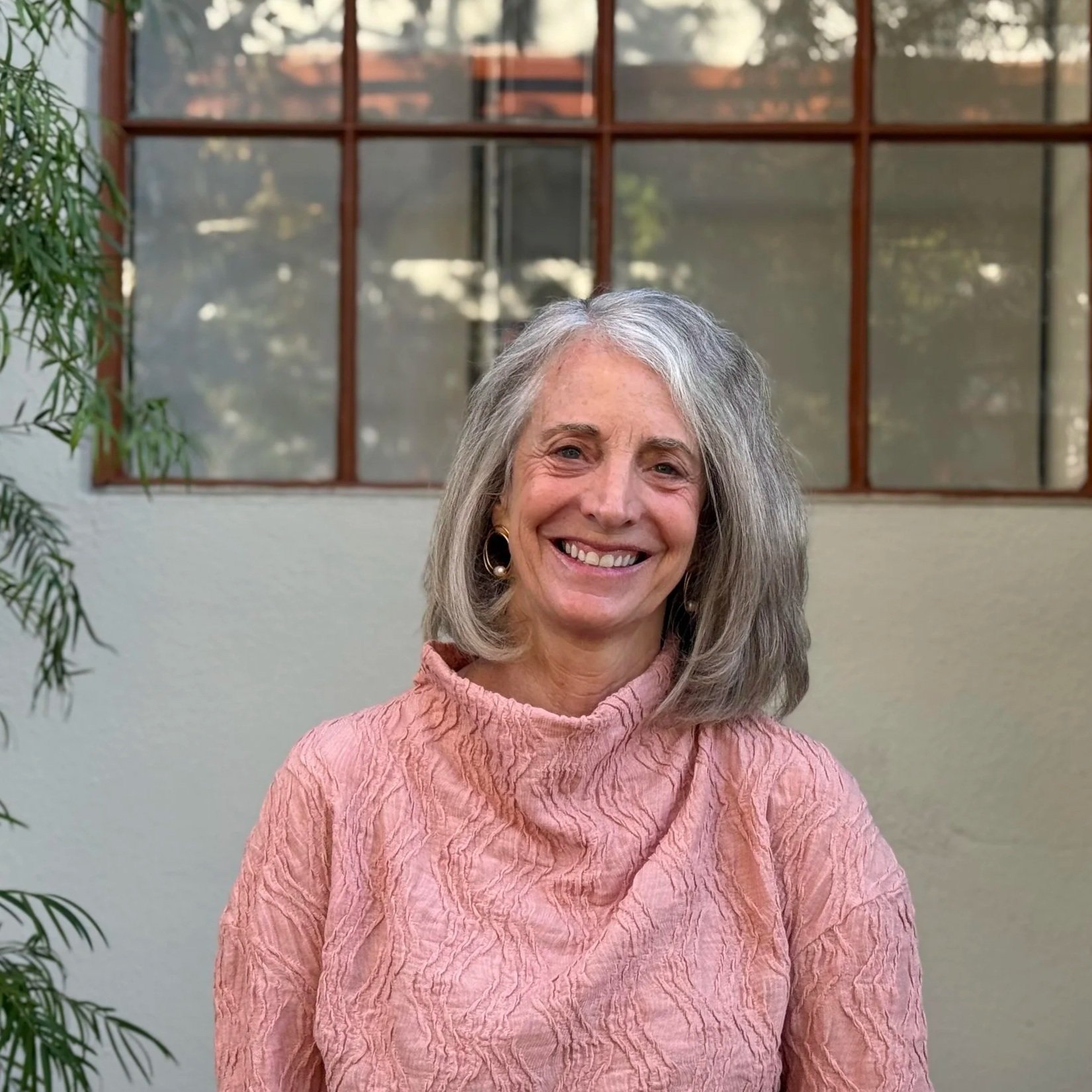 Smiling older woman with gray hair wearing a pink textured top, standing indoors near a window with a plant on the side.