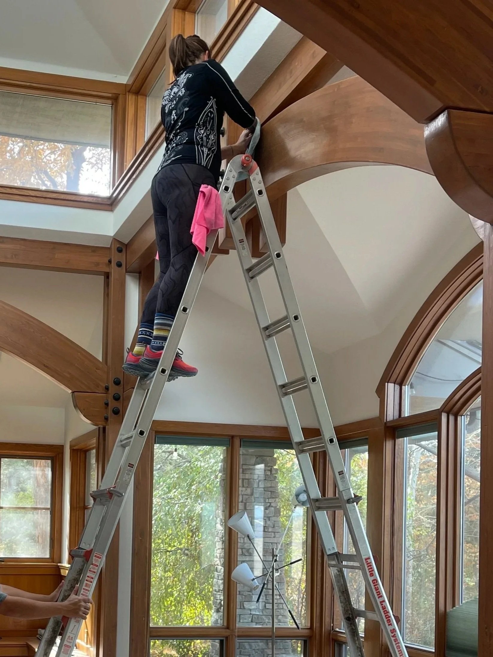 A person standing on a ladder cleaning a large wooden chandelier in a room with large windows and abundant natural light.