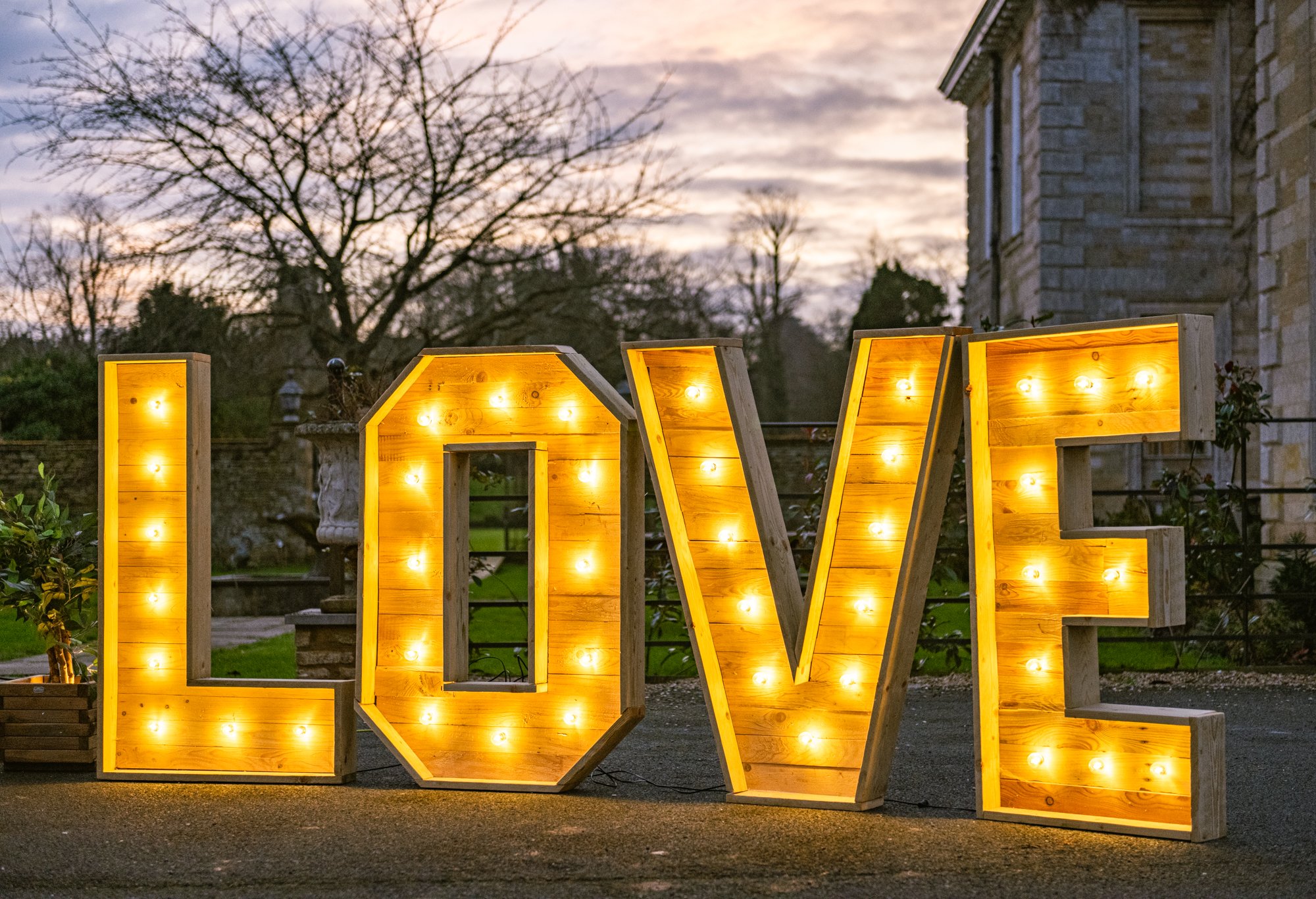Large wooden letters spelling "LOVE" with lights, set outdoors at dusk, trees in the background.