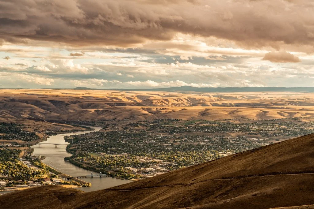 Aerial view of a river winding through a town with bridges, surrounded by rolling hills and mountains under a cloudy sky.