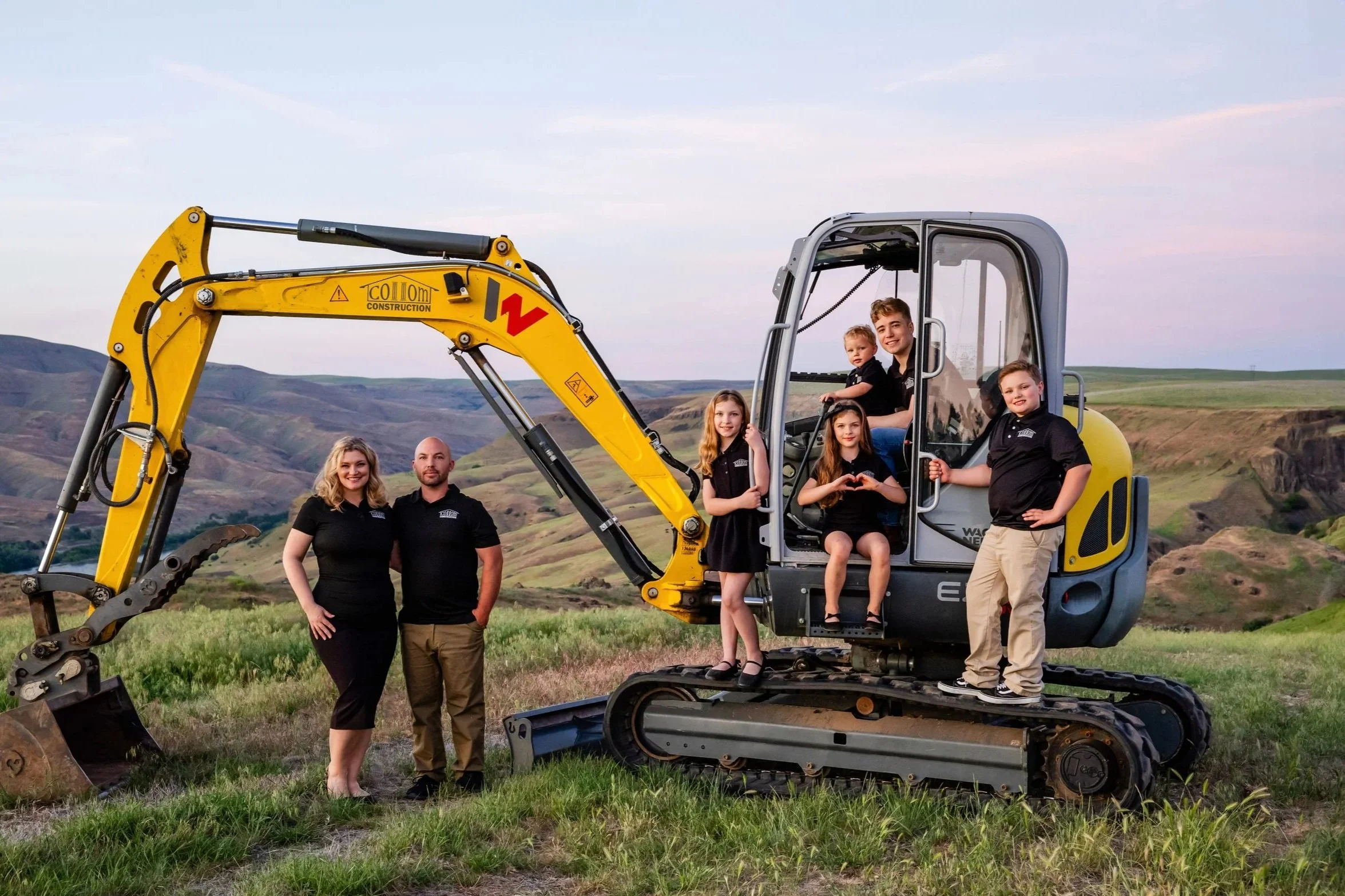 A group of six people, including two adults and four children, standing around a small yellow excavator on a grassy hillside with rolling hills in the background during sunset.