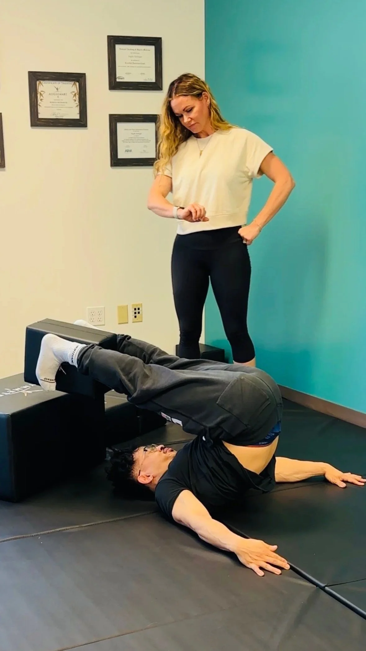 A woman standing and looking down at a man lying on a mat with his legs elevated on exercise blocks, depicted during a corrective exercise training session.